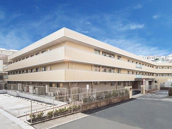 ベージュの外壁の３階建ての建物がある敷地内には駐車場があり、駐車場のそばには草木が植えられている。