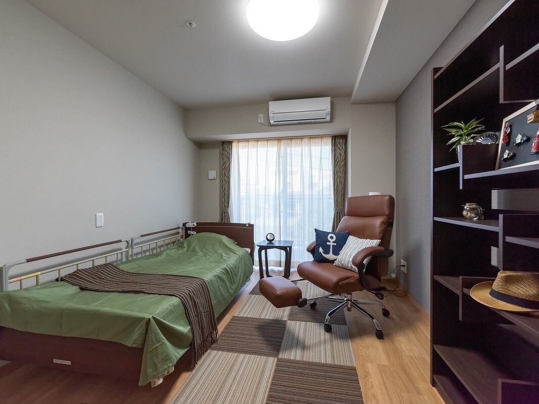 エアコンが付いた部屋には寝具付きのベッドが置かれ、大きな棚や窓があり、ベッドサイドには緊急通報装置がある。