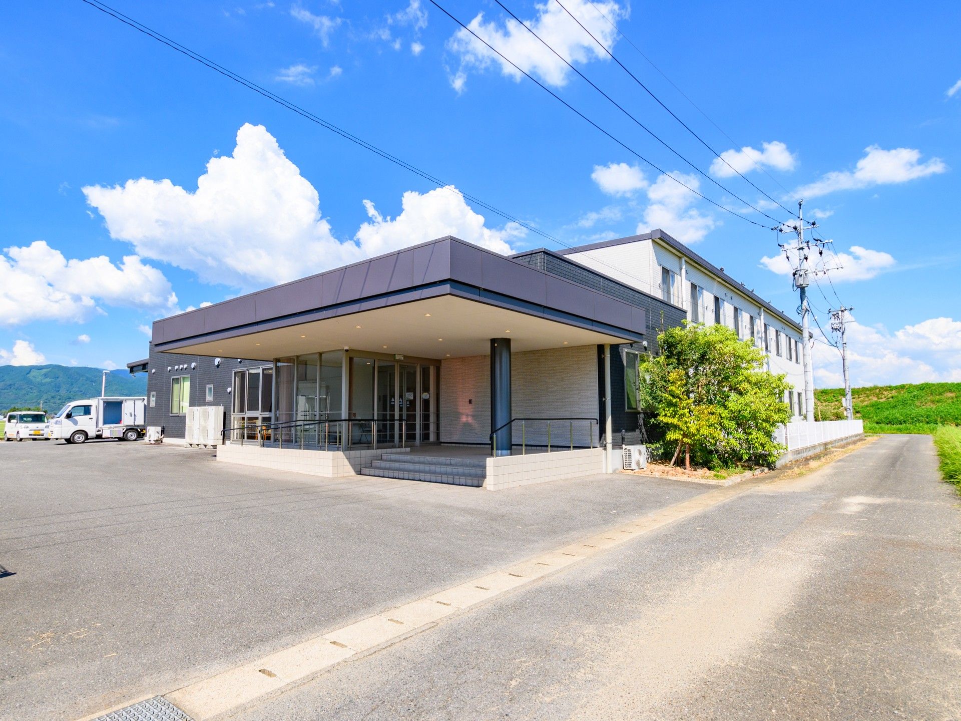 白を基調とした2階建ての建物には黒い屋根が付いた玄関があり、玄関部分は階段と手すり付きのスロープがあり、木も植えられている。