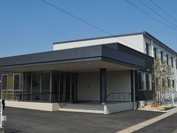 モノトーンの落ち着いた雰囲気の外観。白を基調とした２階建ての建物で、周辺には木が植えられ、駐車場もある。