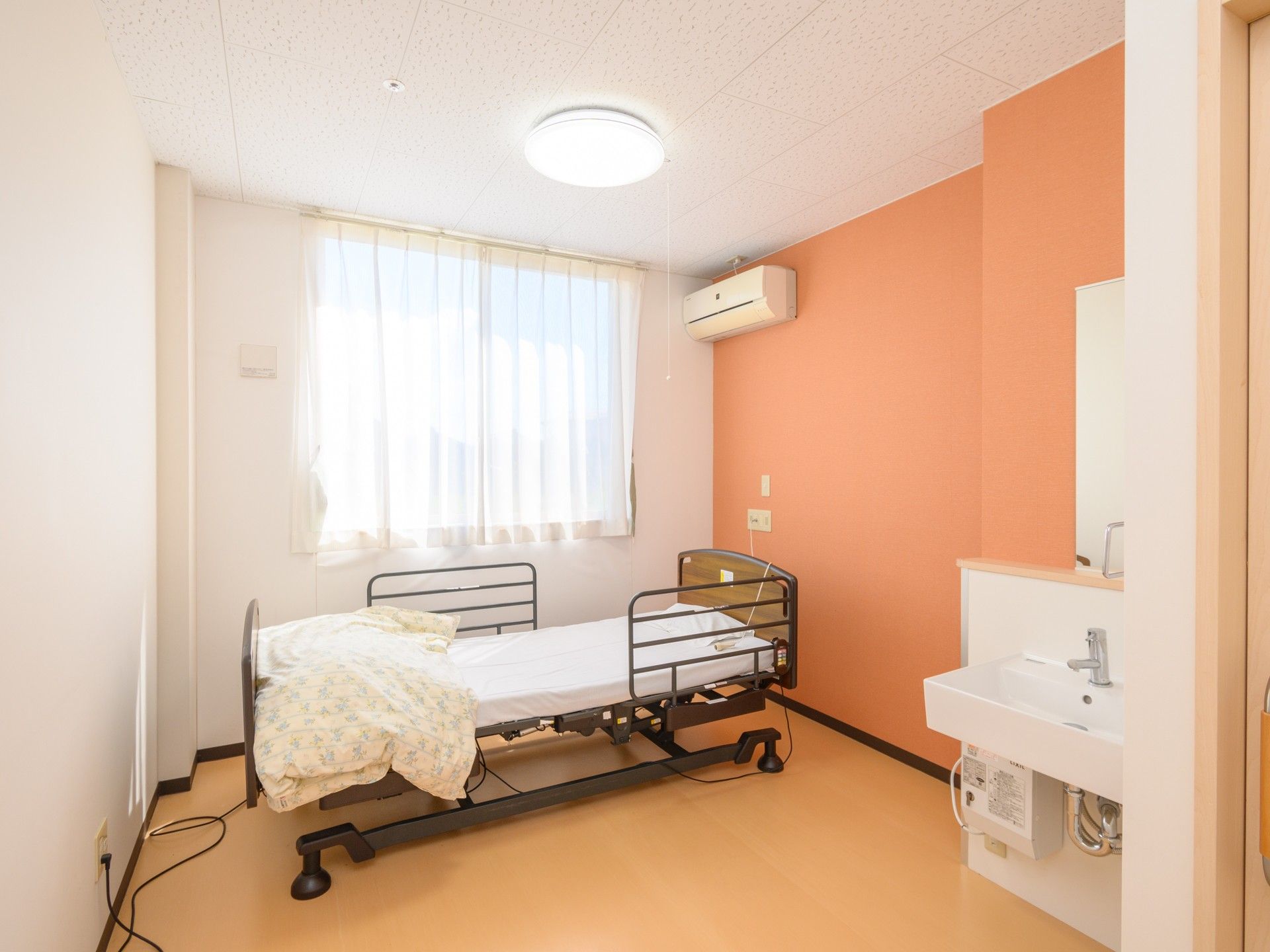 寝具付きのベッドやテーブルなどが設置された部屋。壁紙は白を基調としているが、一面がモスグリーン。窓にはカーテンが付いている。
