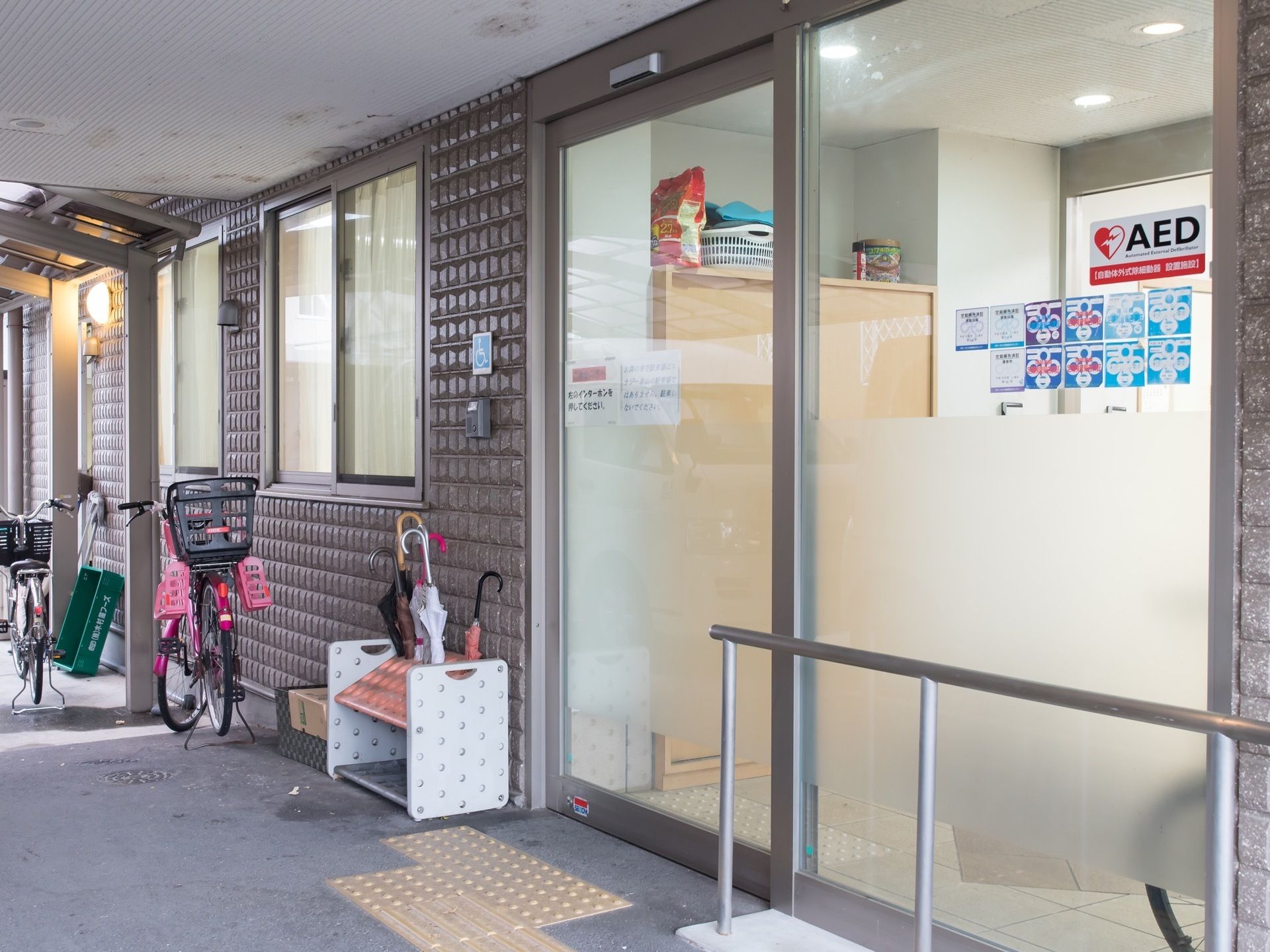 ガラスサッシの自動扉が設置された施設入口である。扉中央はぼかしが入り、見えすぎない工夫がされている。足元には点字ブロックが配備されている。