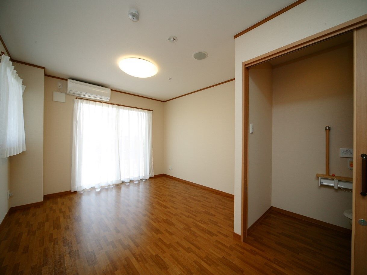 車いす対応の広いトイレが付いたフローリングの部屋にはレースカーテンとカーテンが付いた大きな窓があり、エアコンも設置されている。