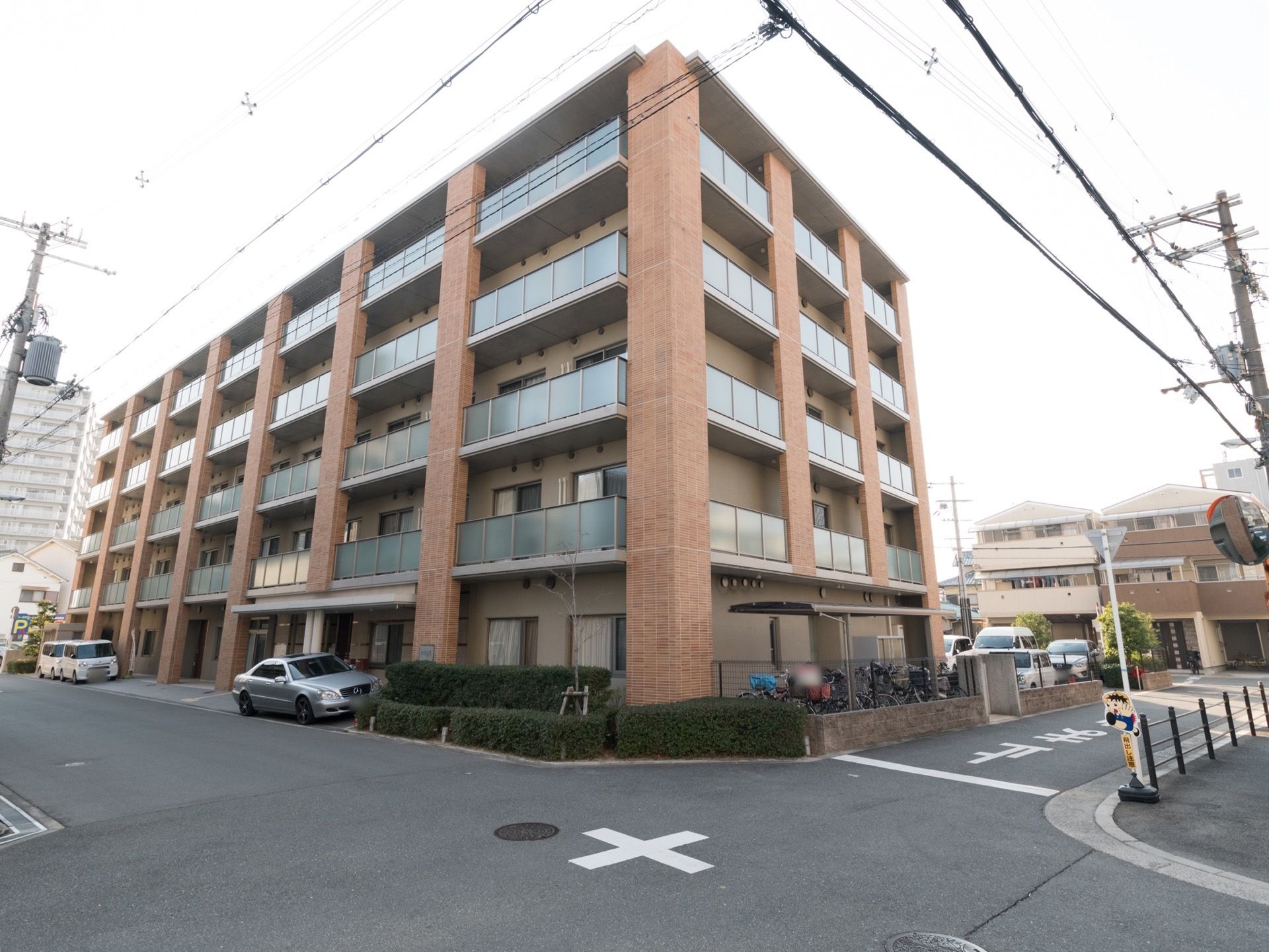 5階建の建物。周囲は住宅街で十字路がある。建物の前にはシルバーの車1台と白いボックスかーが2台停まっている。