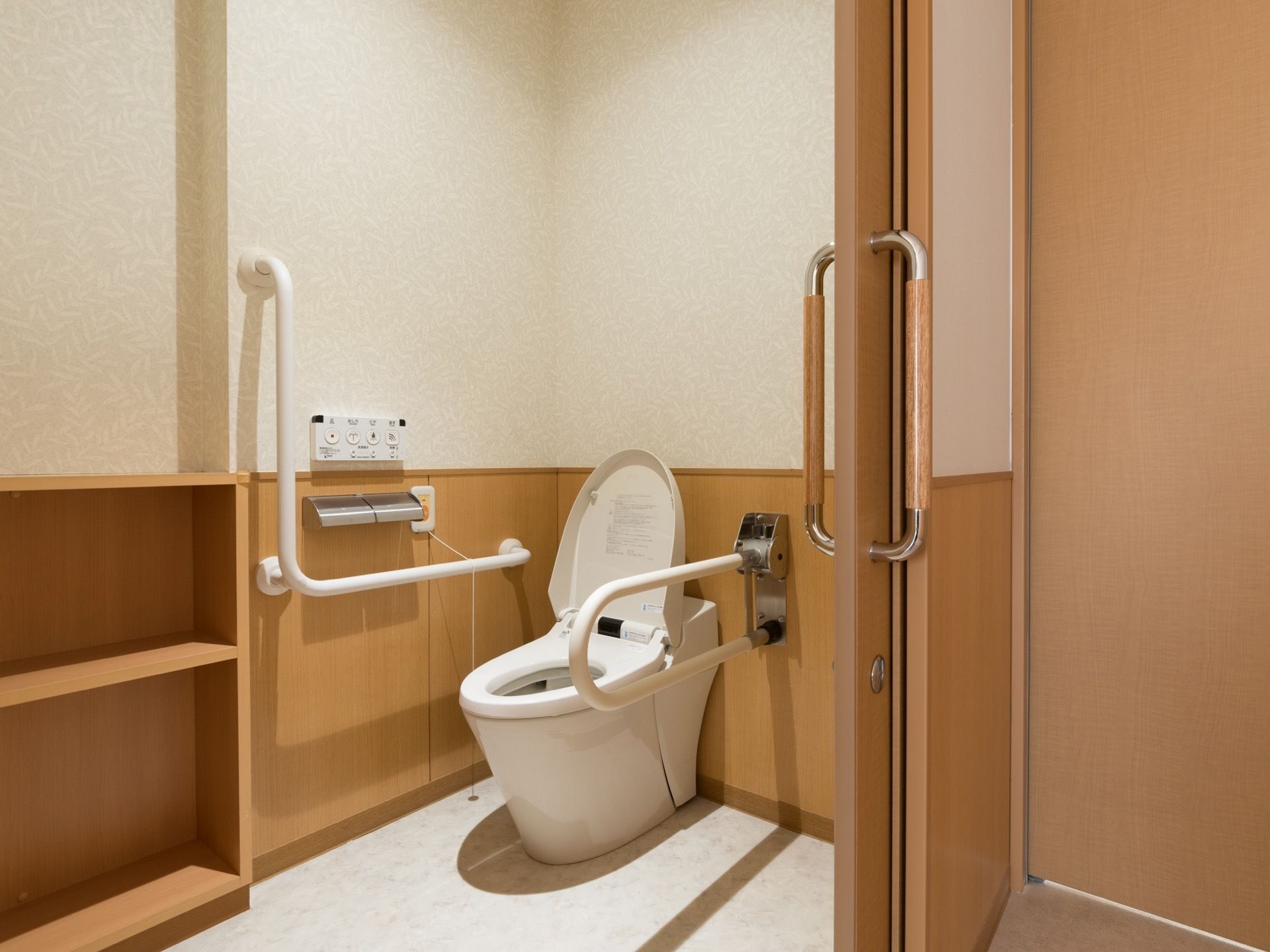 広いトイレは温水洗浄機能付き。室内には2段の棚がある。トイレの両脇には手すりがついている。扉は引き戸。