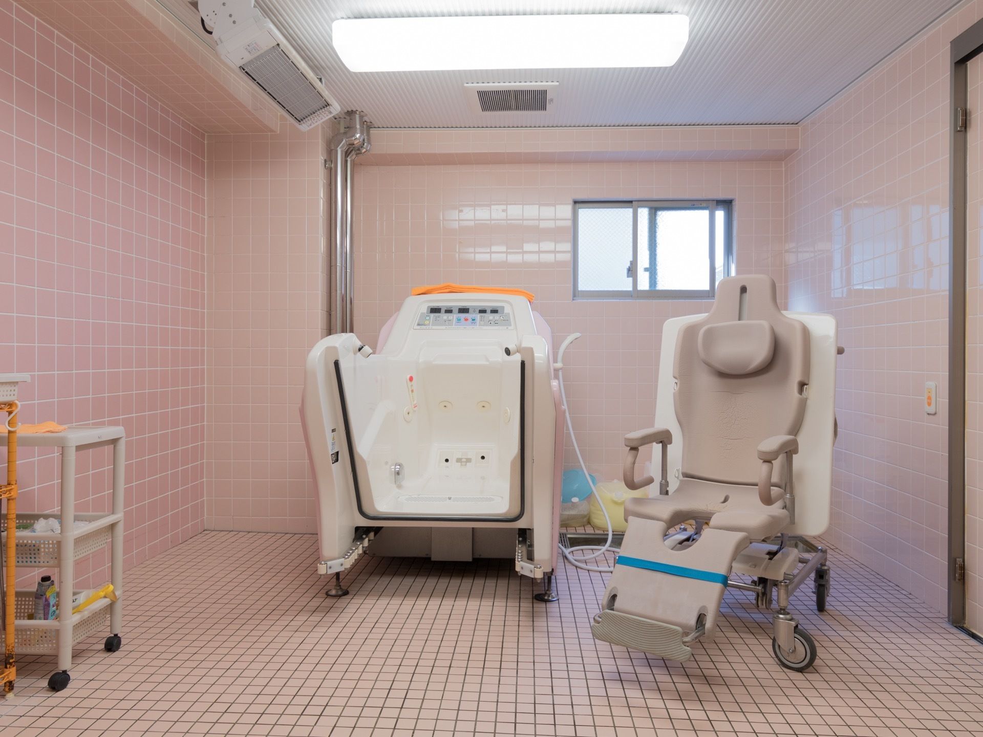 座ったまま使用できる介護浴槽がピンクのタイル貼りの部屋に置かれている。2段の棚にはスポンジやスプレーが置かれている。