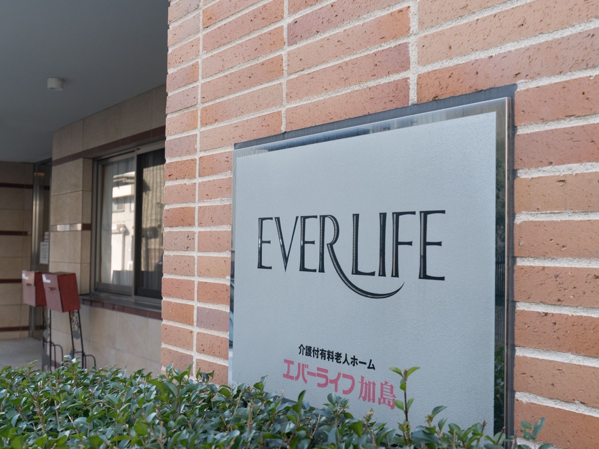 壁はレンガ造りで施設名の銀色のプレートが貼られ、その前に植え込みがある。左手奥にはガラス戸と出入口が見える。