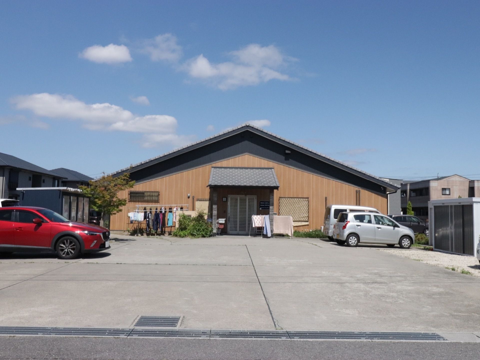 施設は三角屋根の１階建ての建物である。施設前では洗濯物や布団が干されている。施設の前は駐車スペースが広がっている。