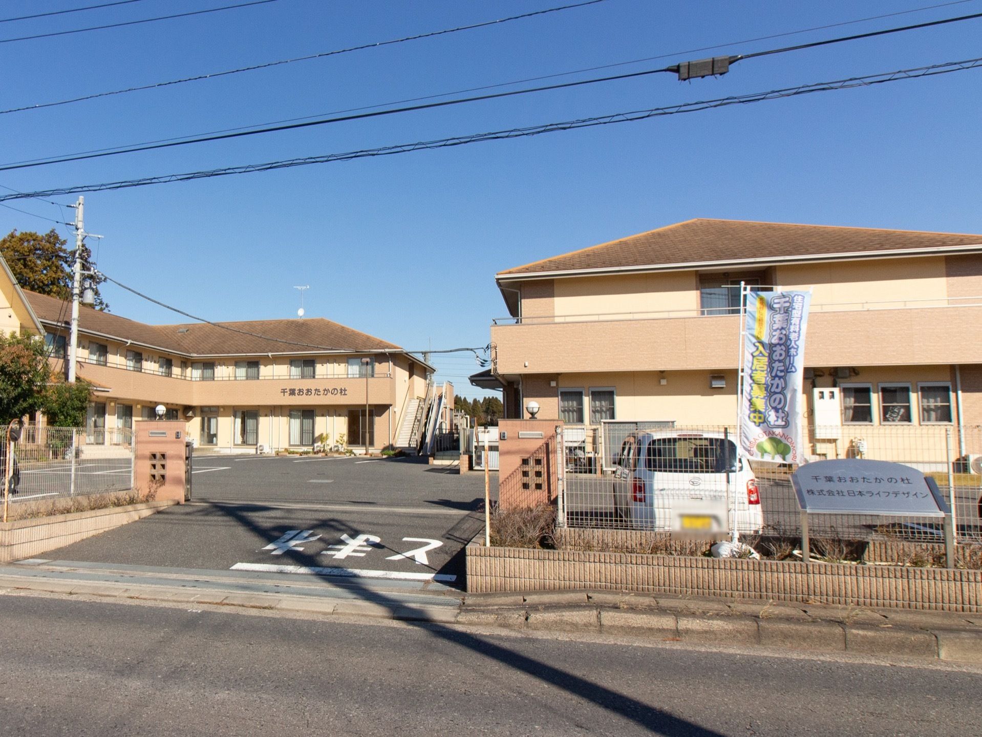 ゲートの奥に２階建ての建物があり、隣には駐車場が完備されている。施設前の道は、歩道が整備されている。