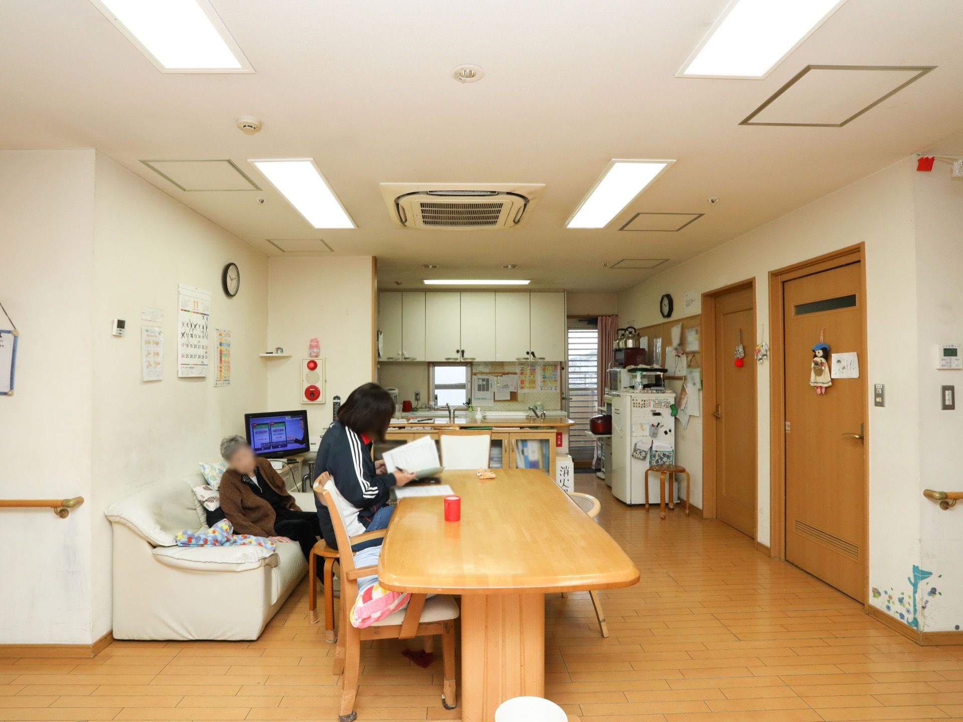 共有スペースにダイニングテーブル、椅子、ソファー、テレビなどが置かれている。ソファーに入居者様が座っている。