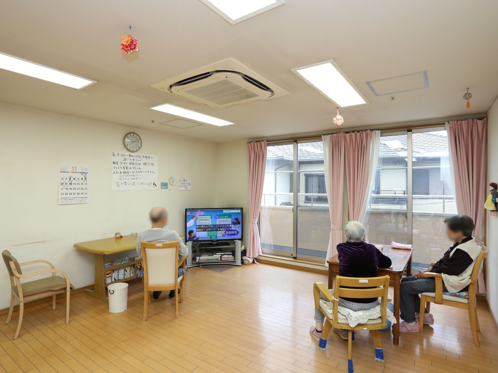 大きな窓が設置された共有スペース。入居者様がダイニングテーブル前の椅子に座り、テレビ鑑賞を楽しんでいる。
