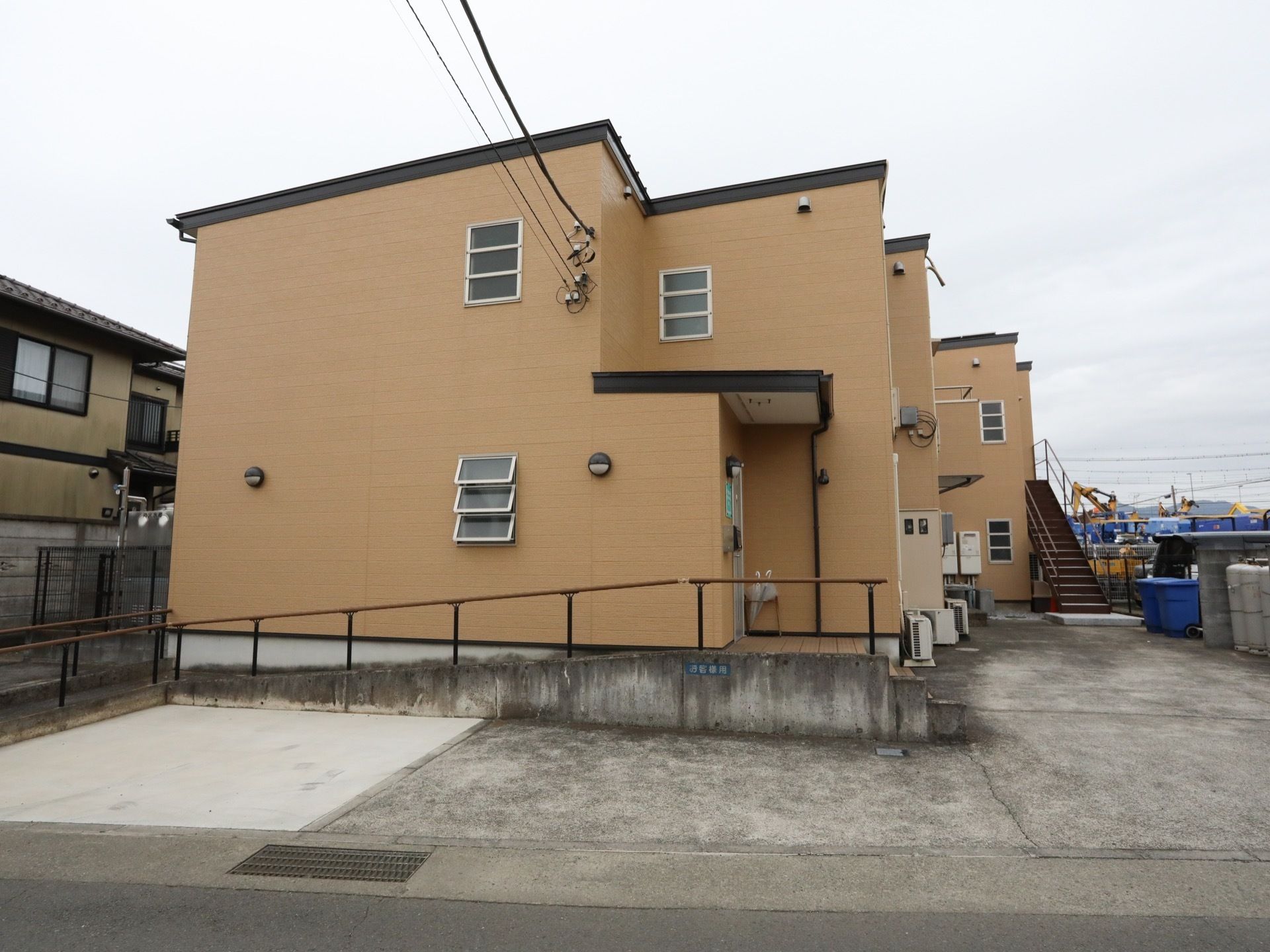 ベージュを基調とした２階建ての施設。施設の敷地内にコンクリートが敷かれている。玄関前には階段とスロープが設置されている。