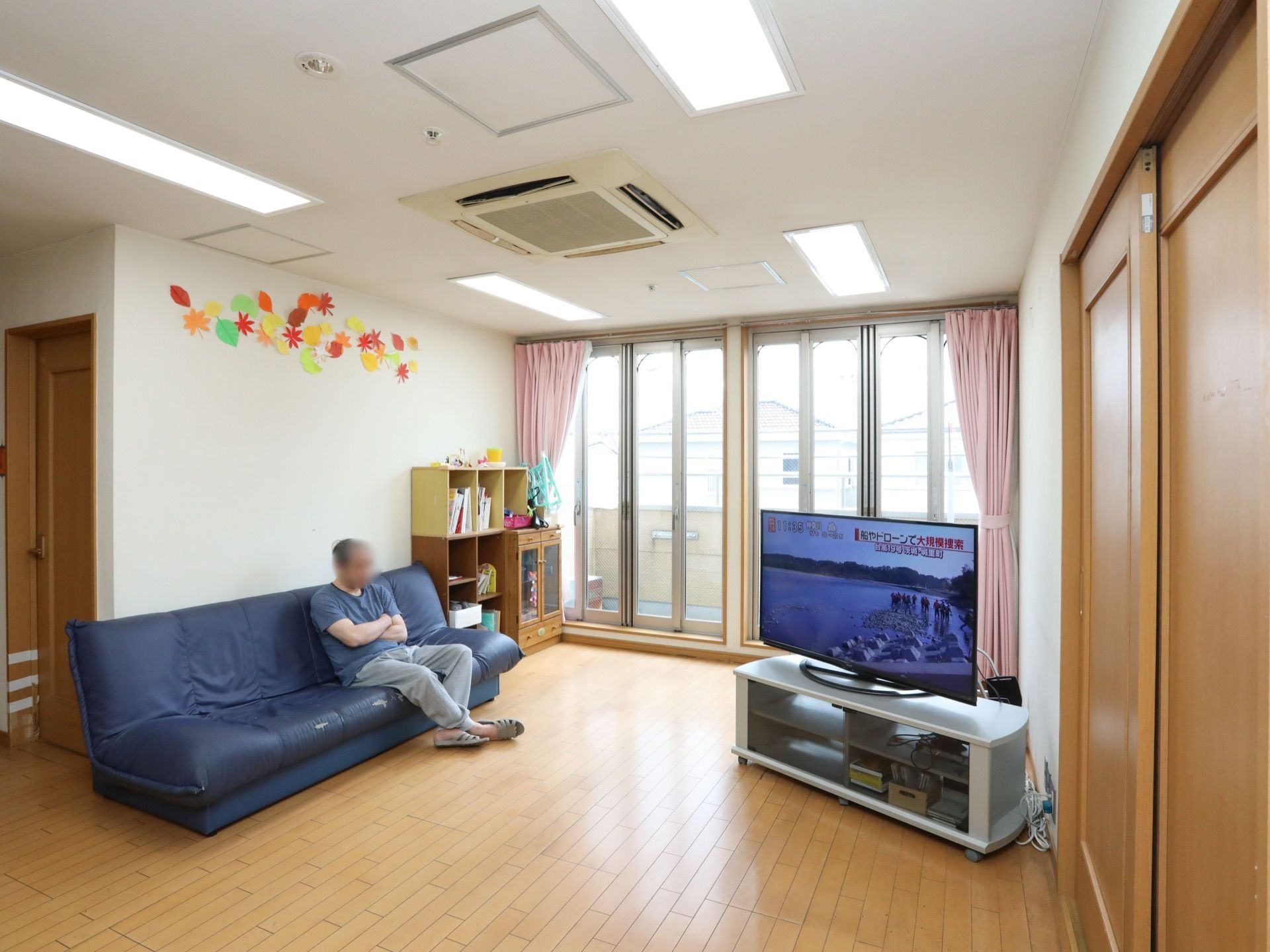 大きな窓があり、日当たりの良いスペースにネイビーのソファーとテレビが設置されている。ソファーの横には本棚があり、本が並べられている。