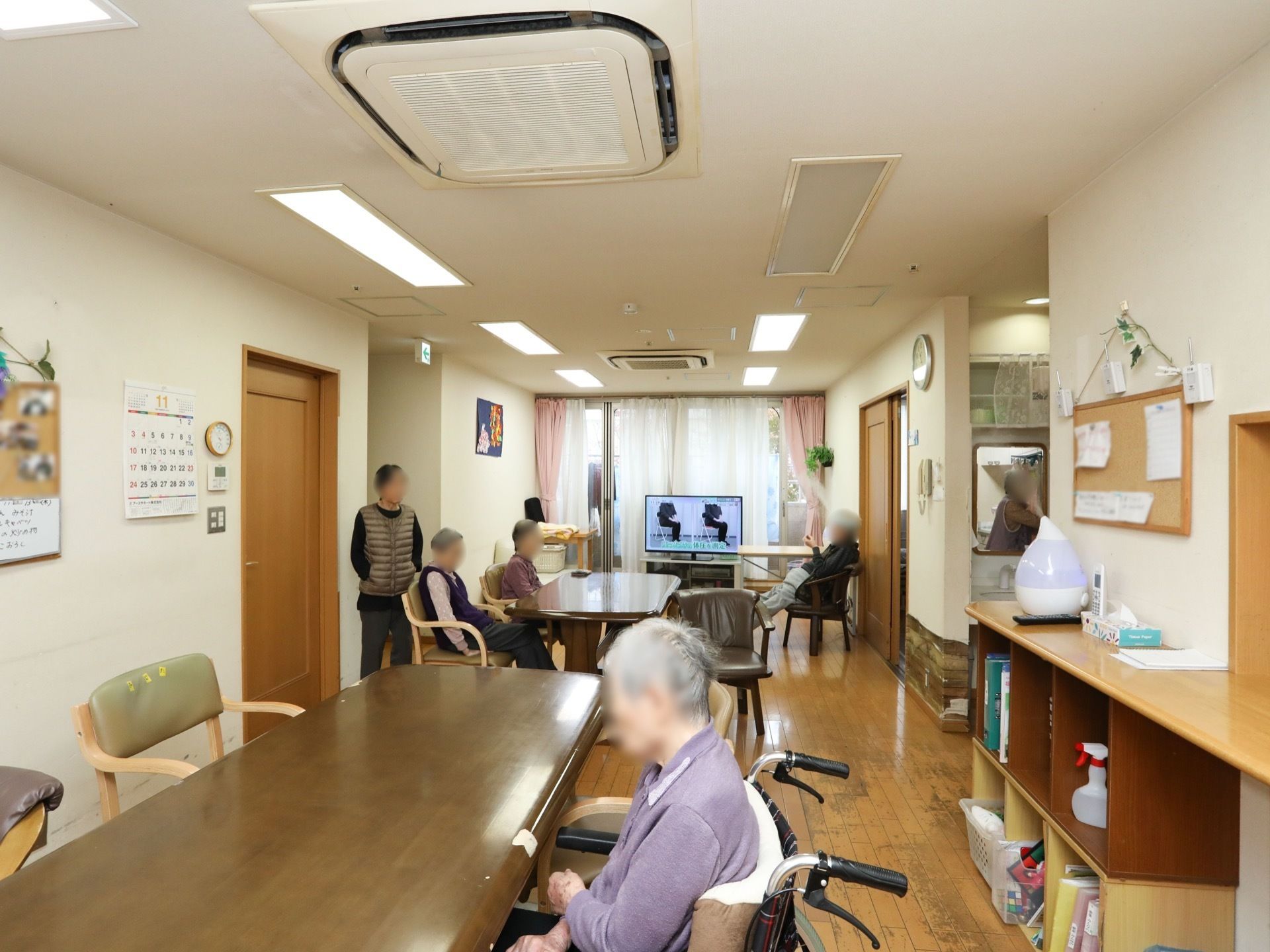 共有スペースのダイニングテーブルを囲んで入居者様が座り、テレビを見たり、くつろいだりしている。壁にカレンダーが掛けられている。