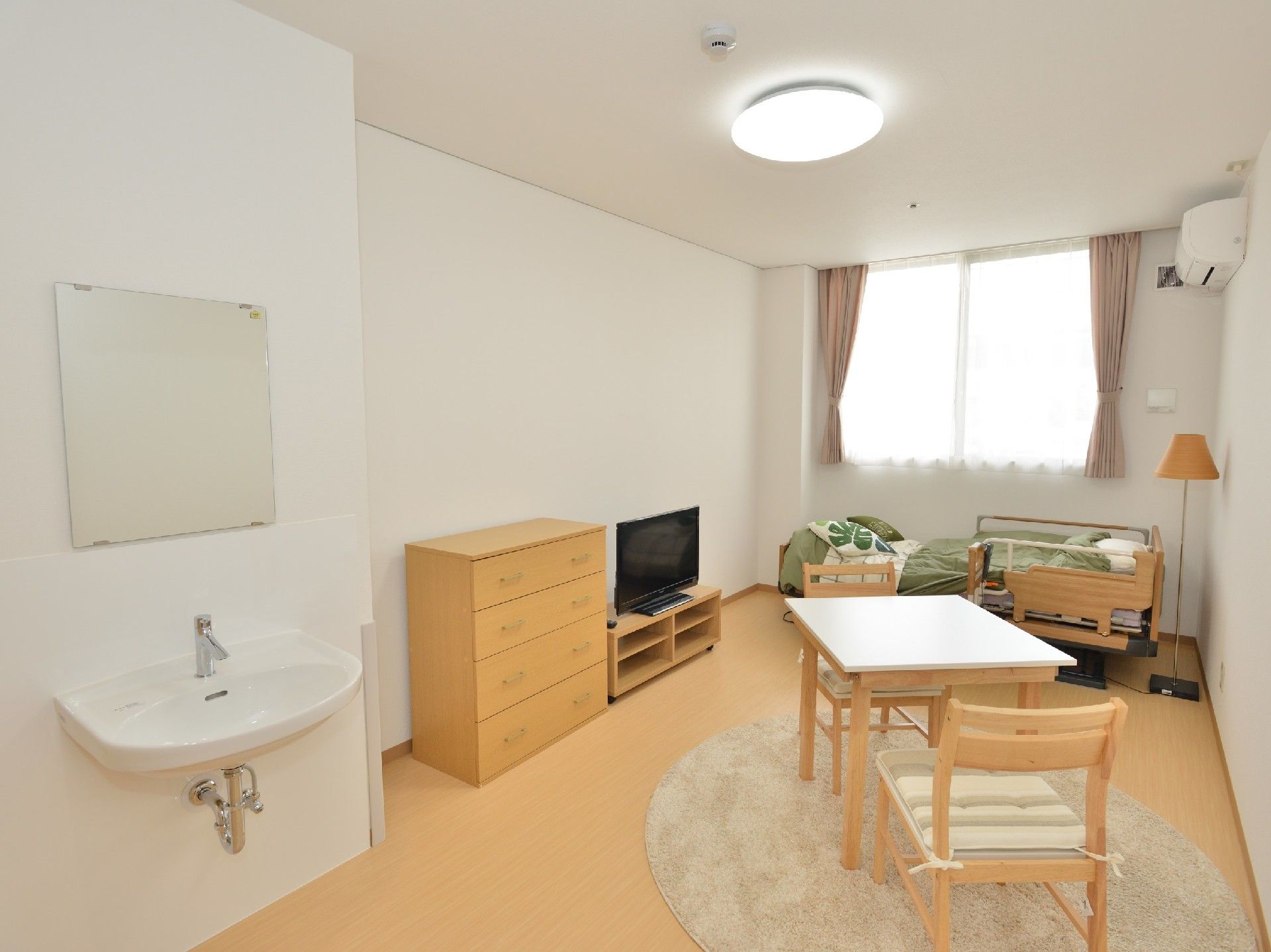 洗面台が手前に写っている小さな居室の写真。モダンなデザイン尾家具類、介護用ベッドが置いてあるスペース