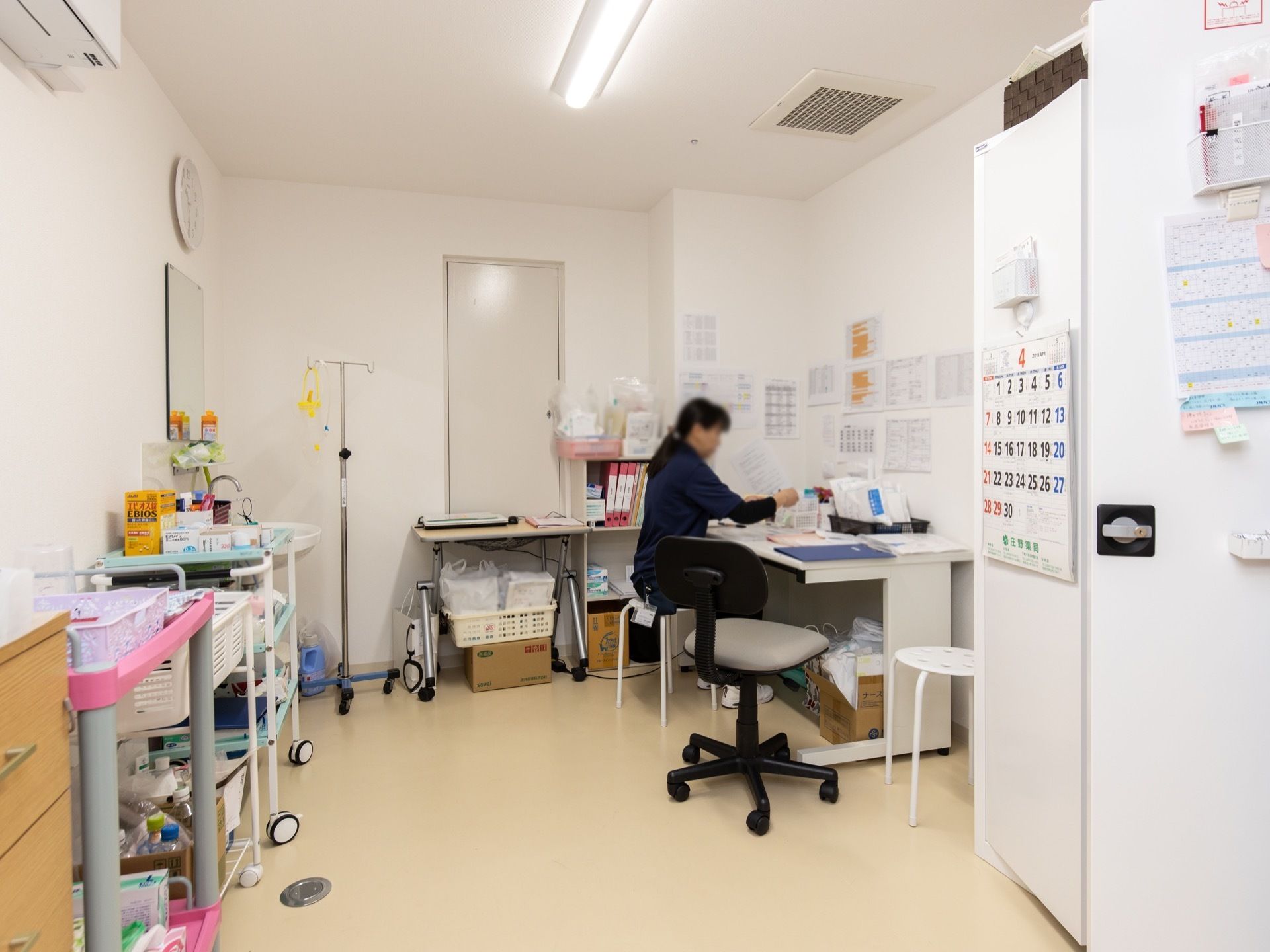 医務室で、女性スタッフが机に向かって作業をしている。可動式のワゴンがいくつかあり、かごにいろいろなボトルや器具が入っている。
