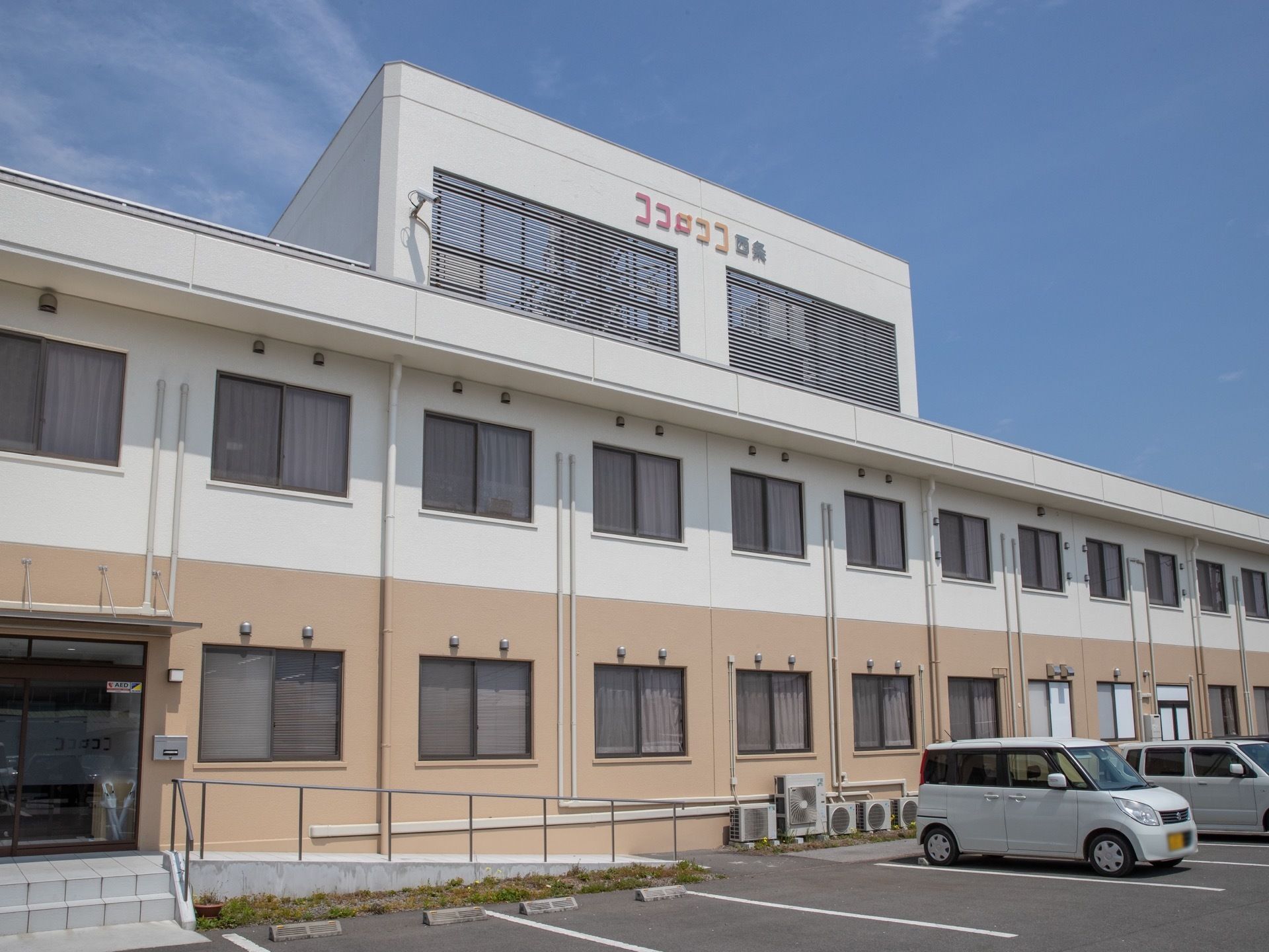 玄関の前には、２段の階段と、ゆるやかなスロープが設置されている。屋上設備の目隠しの外壁に施設名称が表示されている。