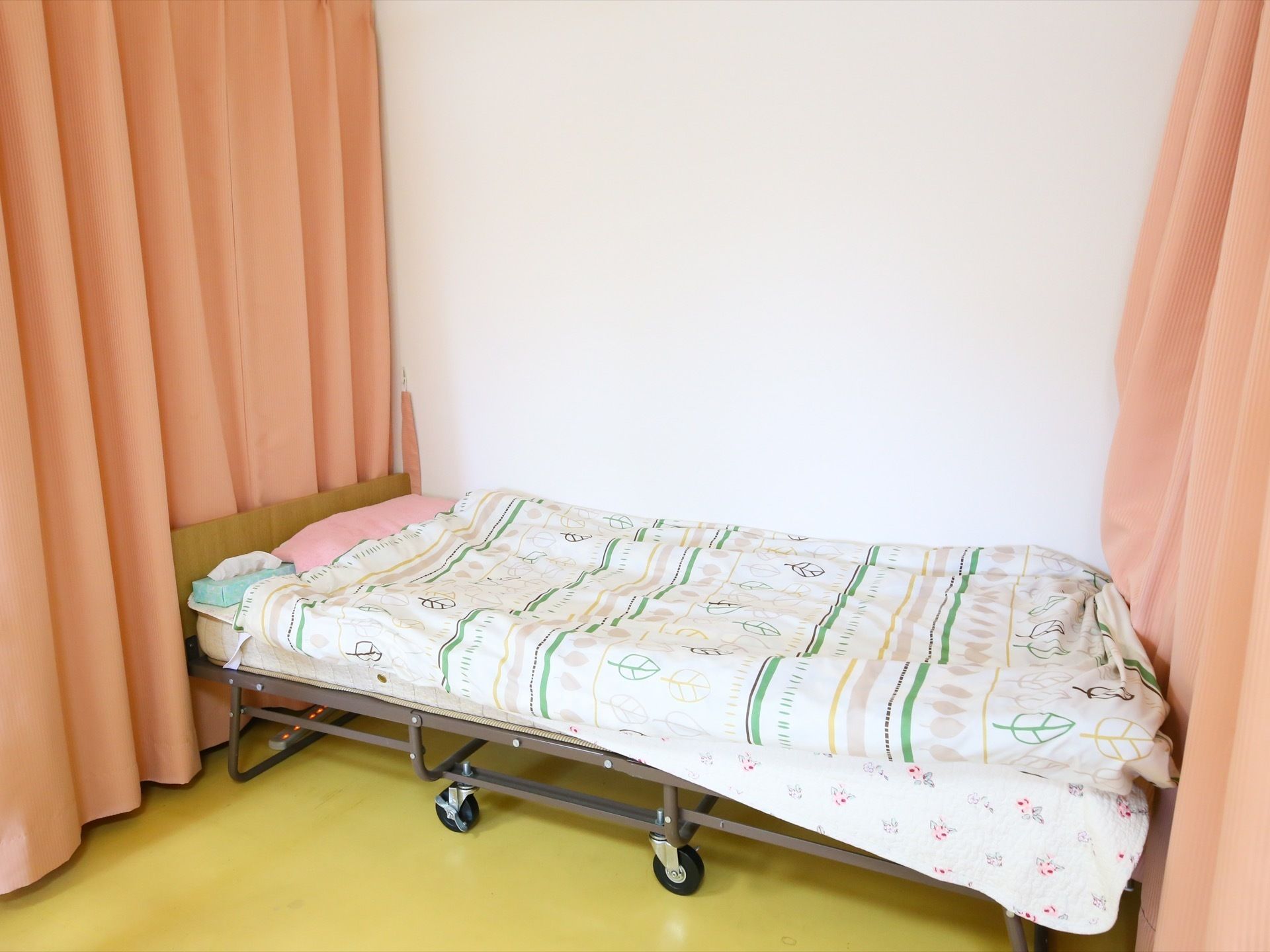 ピンク色の仕切りカーテンが、両側に備えつけられている。キャスター付きの介護専用ベッドが完備されてある。