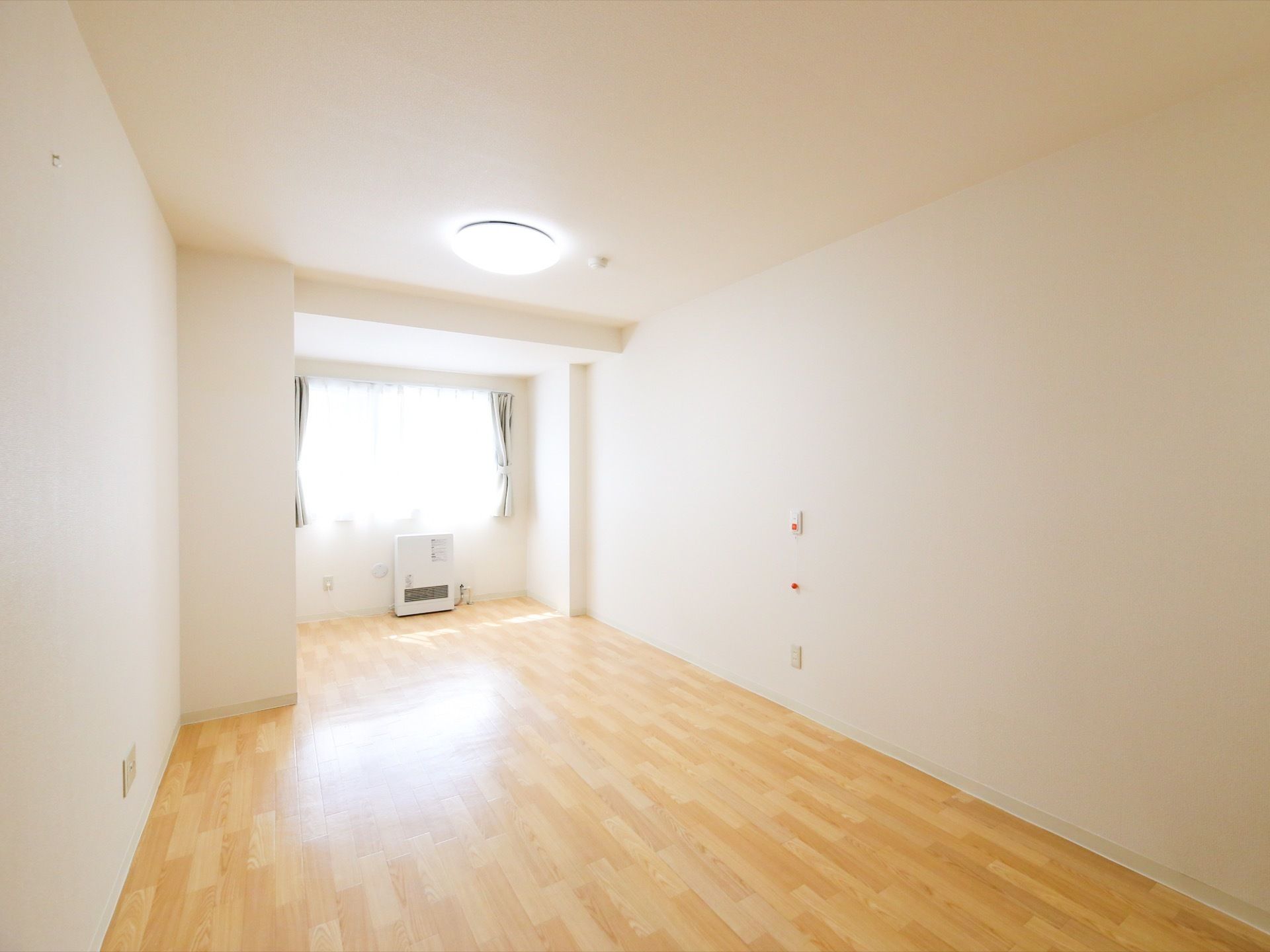 大きな窓から明るい光の差し込む居室の壁は白である。窓際に床置き型の暖房機器が置かれ、壁にはナースコールがある。