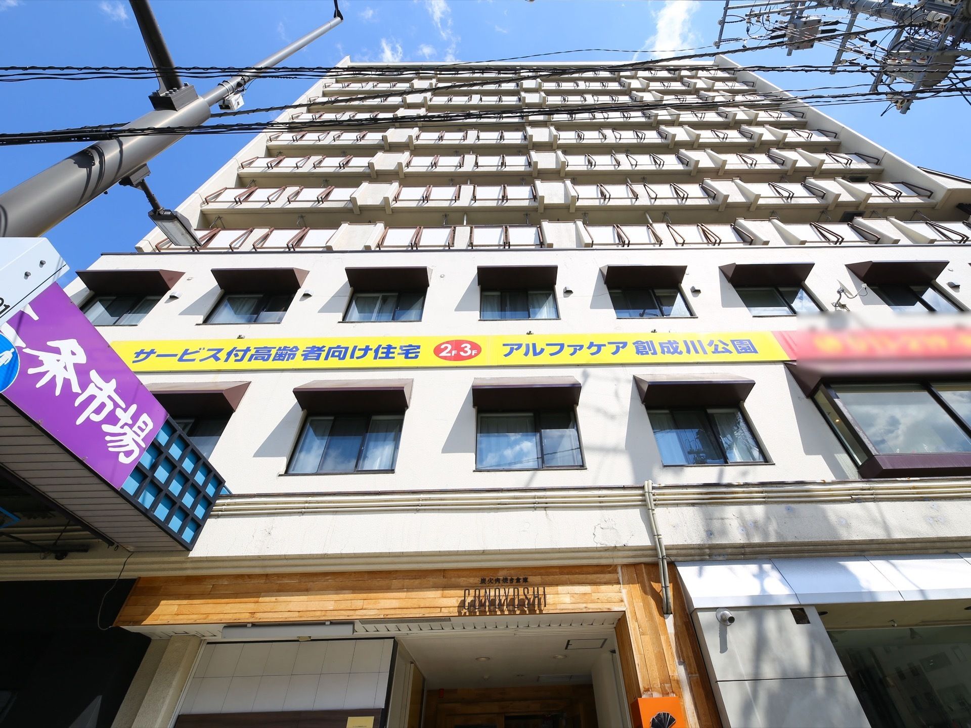 鉄骨の白い建物は11階建で、２階と３階の間に施設の看板がある。建物脇には市場へのアーケード入口が見える。