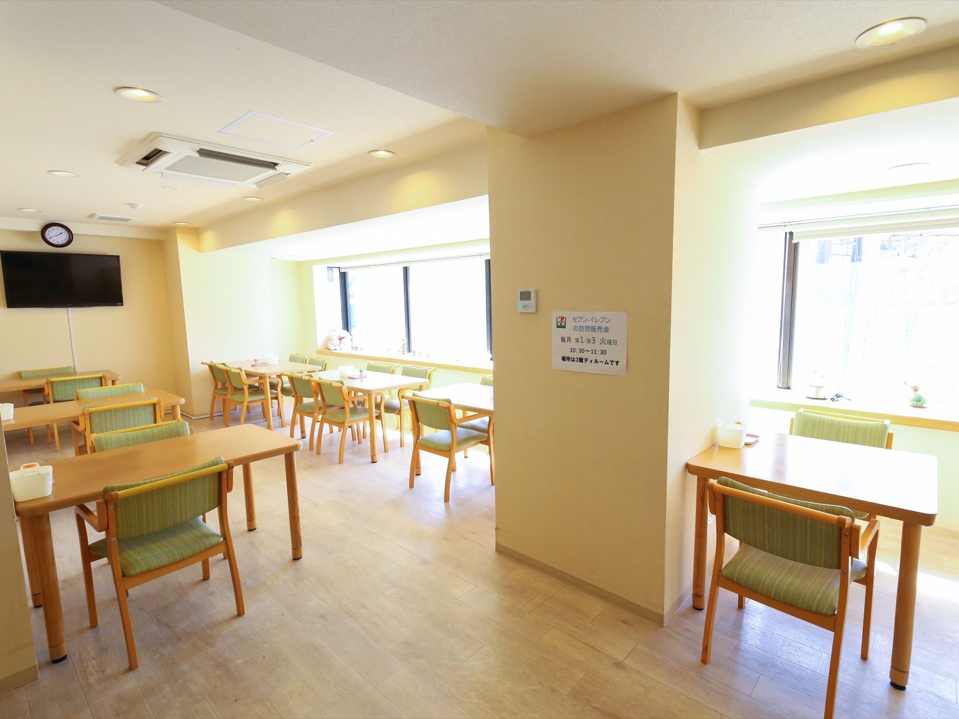 明るい木目調のテーブルにグリーン系の椅子が配置された食堂である。窓から陽射しが入り込み、テレビや時計が壁に設置されている。