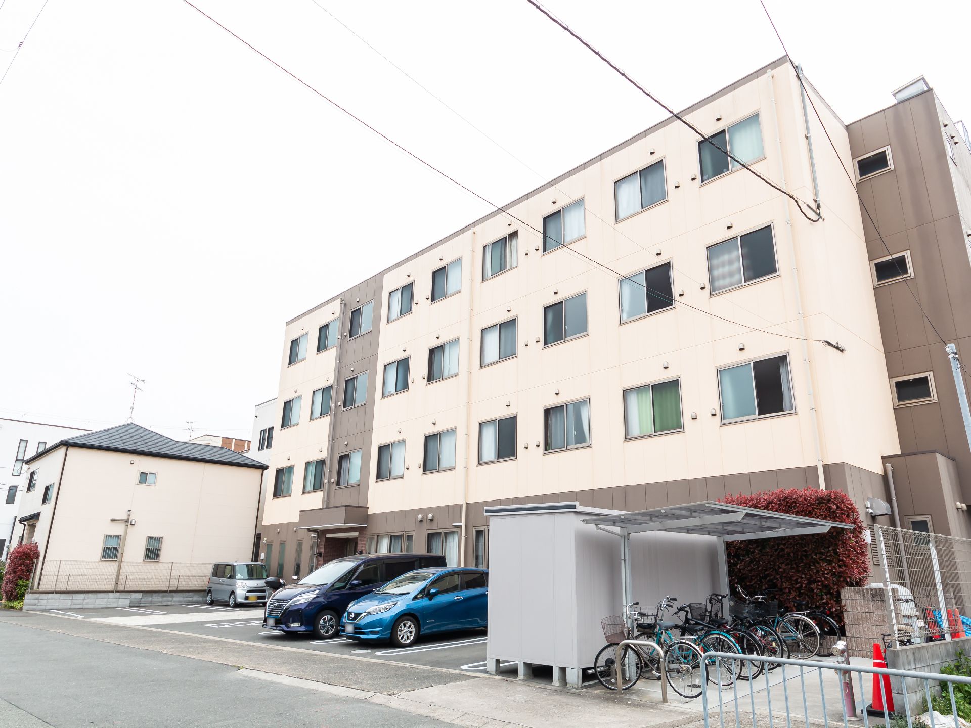 ベージュを基調とした4階建ての建物は窓がたくさんついている。建物の前には駐車スペースもある。