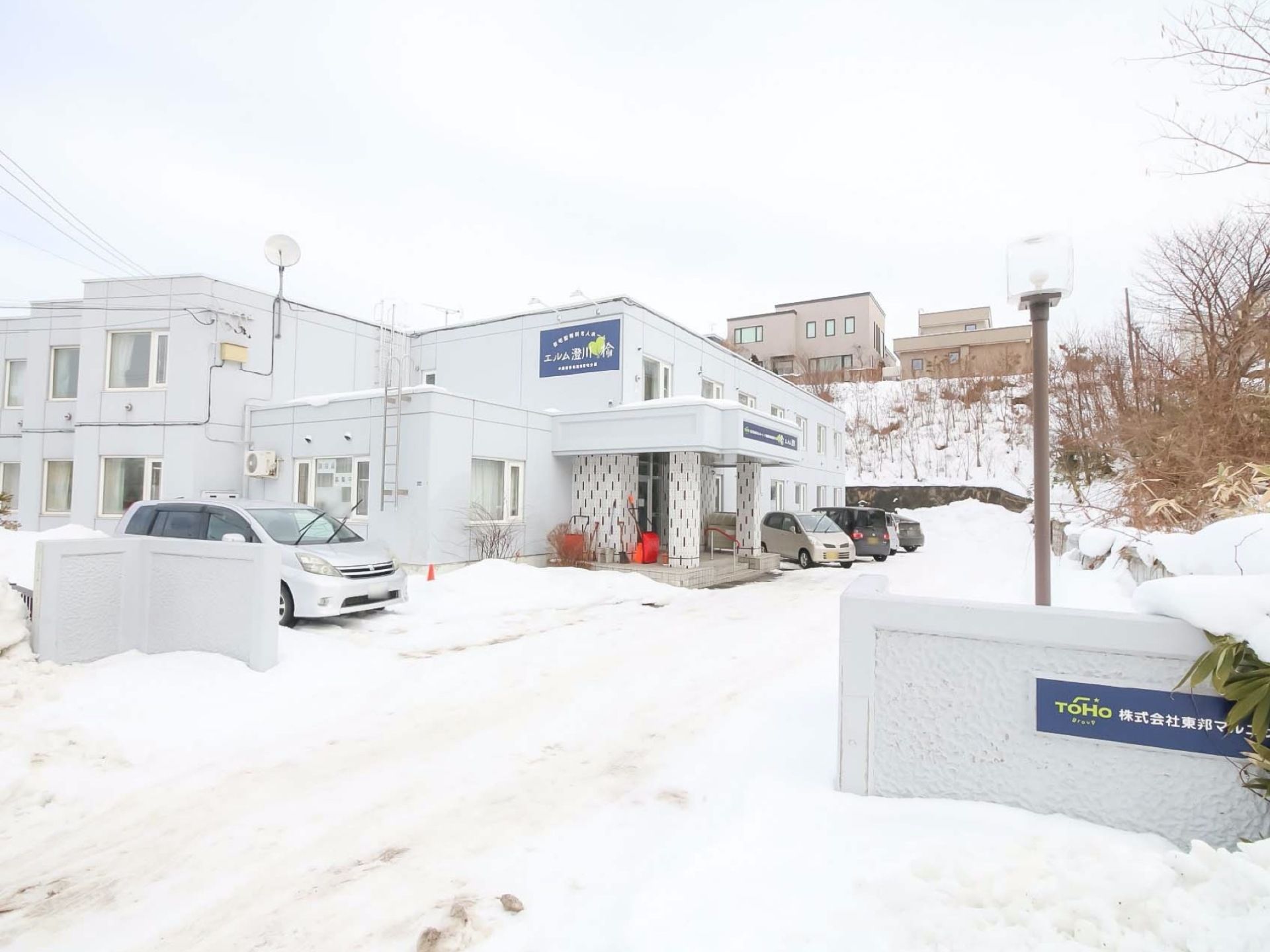 白い２階建ての建物が雪の中に建っている。左側が個室の並ぶフロアになっている。玄関前は、駐車スペースが広く取られている。