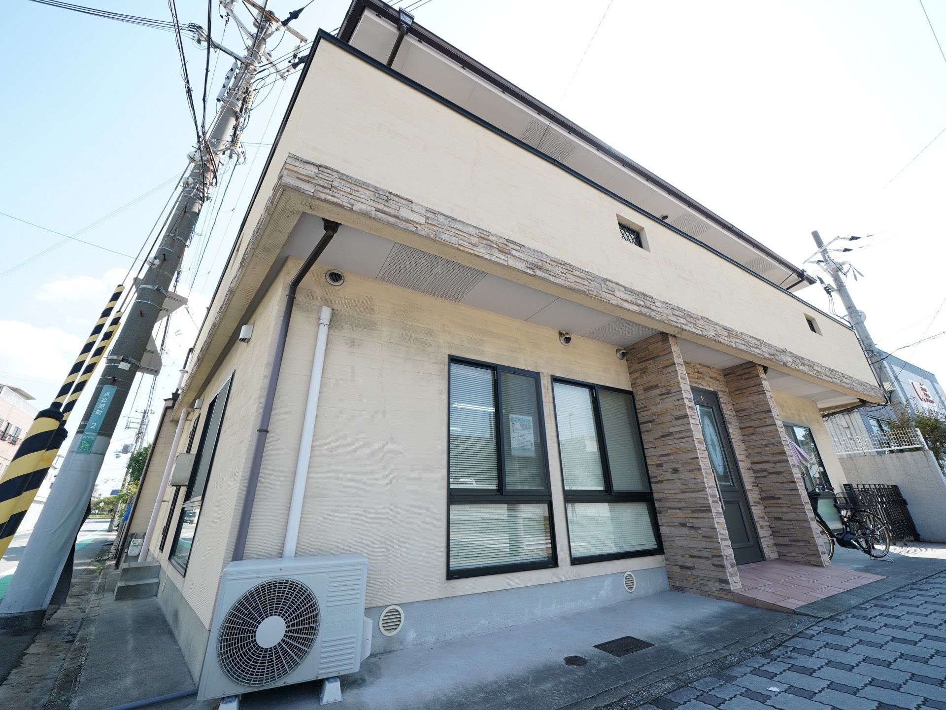 角地に建つ、ベージュ色の外壁の建物である。外にはエアコンの室外機が取りつけてある。玄関の扉はモダンなデザインとなっている。