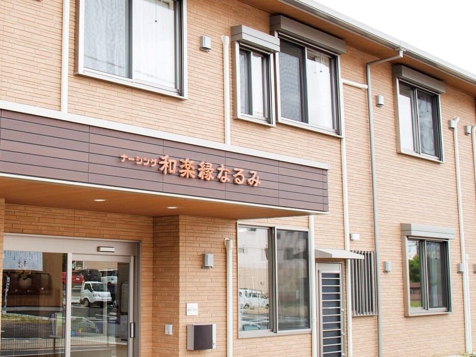 オレンジの文字でホーム名が書かれている屋根が玄関についた２階建ての建物はブラウン系の外壁。