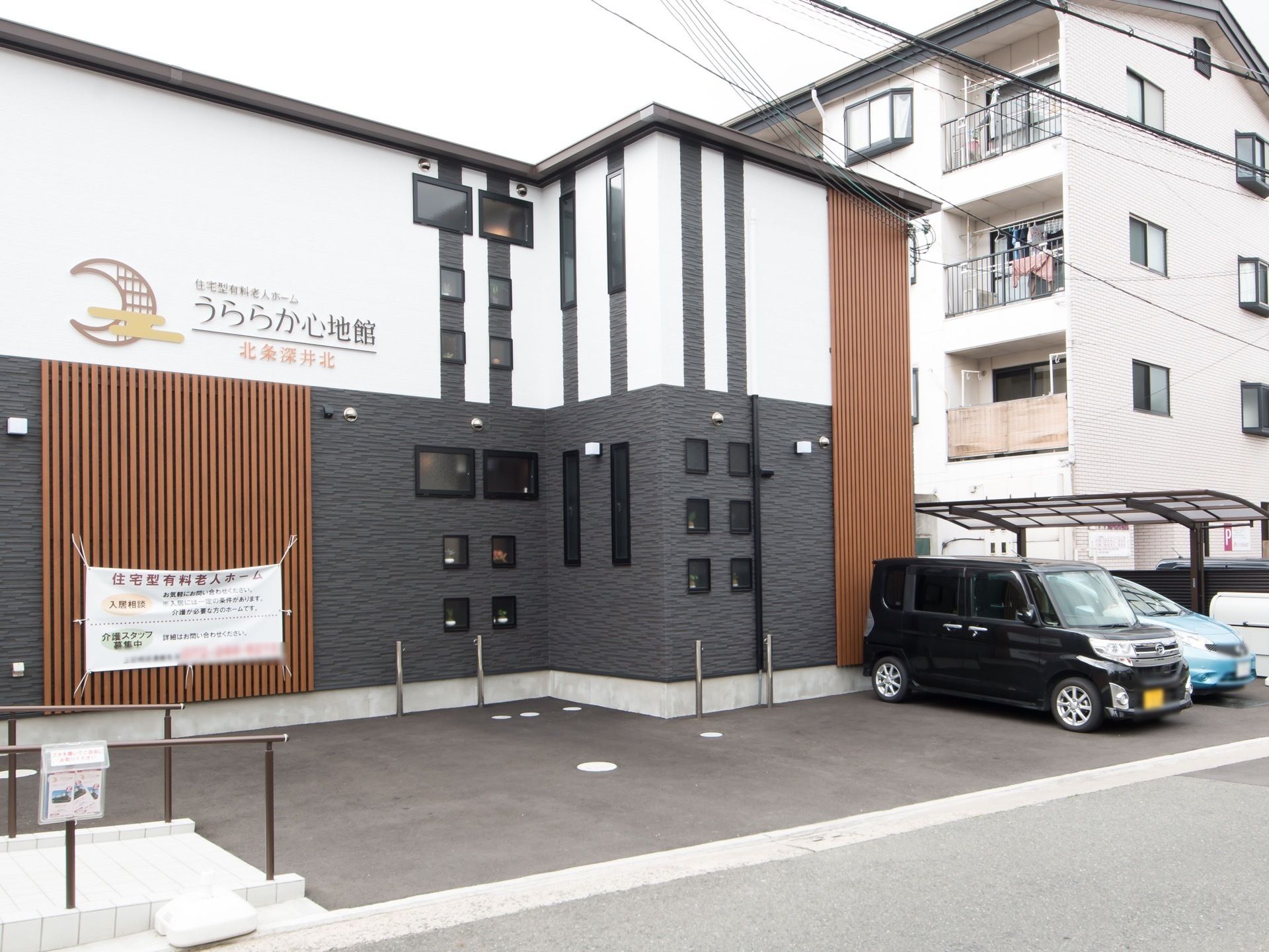 アパートの隣に建つ２階建ての施設。外壁の高い位置に施設名称が明記されており、その下方には施設案内やスタッフ募集を記した幕が掲示されている。