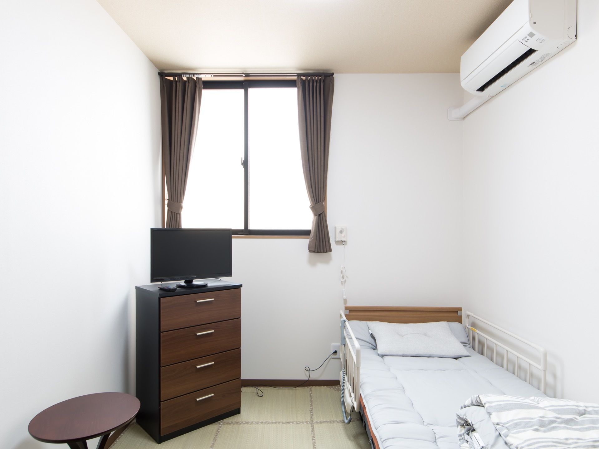部屋の左側の隅に、引き出しが４段ついた収納家具が置かれており、その上に液晶モニターがある。右側にはベッドが据えられている。