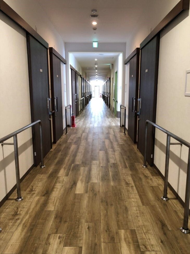 手すりが両サイドに付いた廊下はまっすぐで、突き当りにある窓から陽の光が差している。居室のドアは黒いスライド式のドアで木材をたくさん使用した館内。