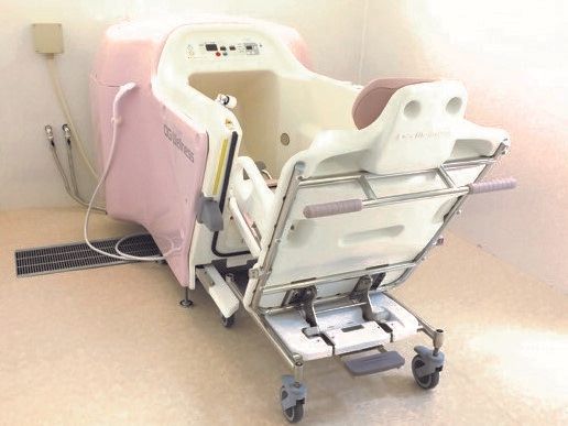 白とピンクの機械浴は車いすのようなデザインの機械に座ったまま入浴可能な設計で、浴槽のような機械にはシャワーも付いている。