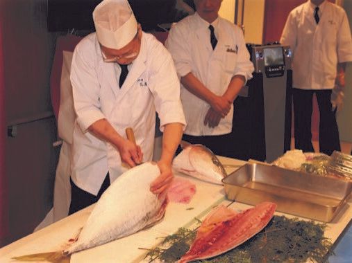大きな魚を包丁一本でさばいている料理人の姿。包丁の上には頭を切断された魚の身があり、半身が海藻の上に置かれている。