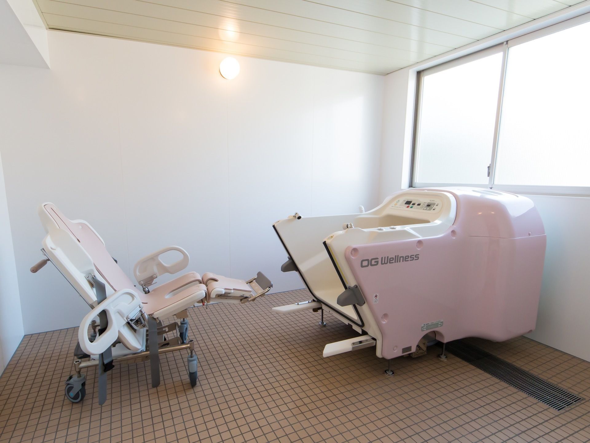 広々とした浴室に機械浴を備えている。側面が開く浴槽の傍に、キャスター付きの椅子がある。大きな窓があり明るい空間である。