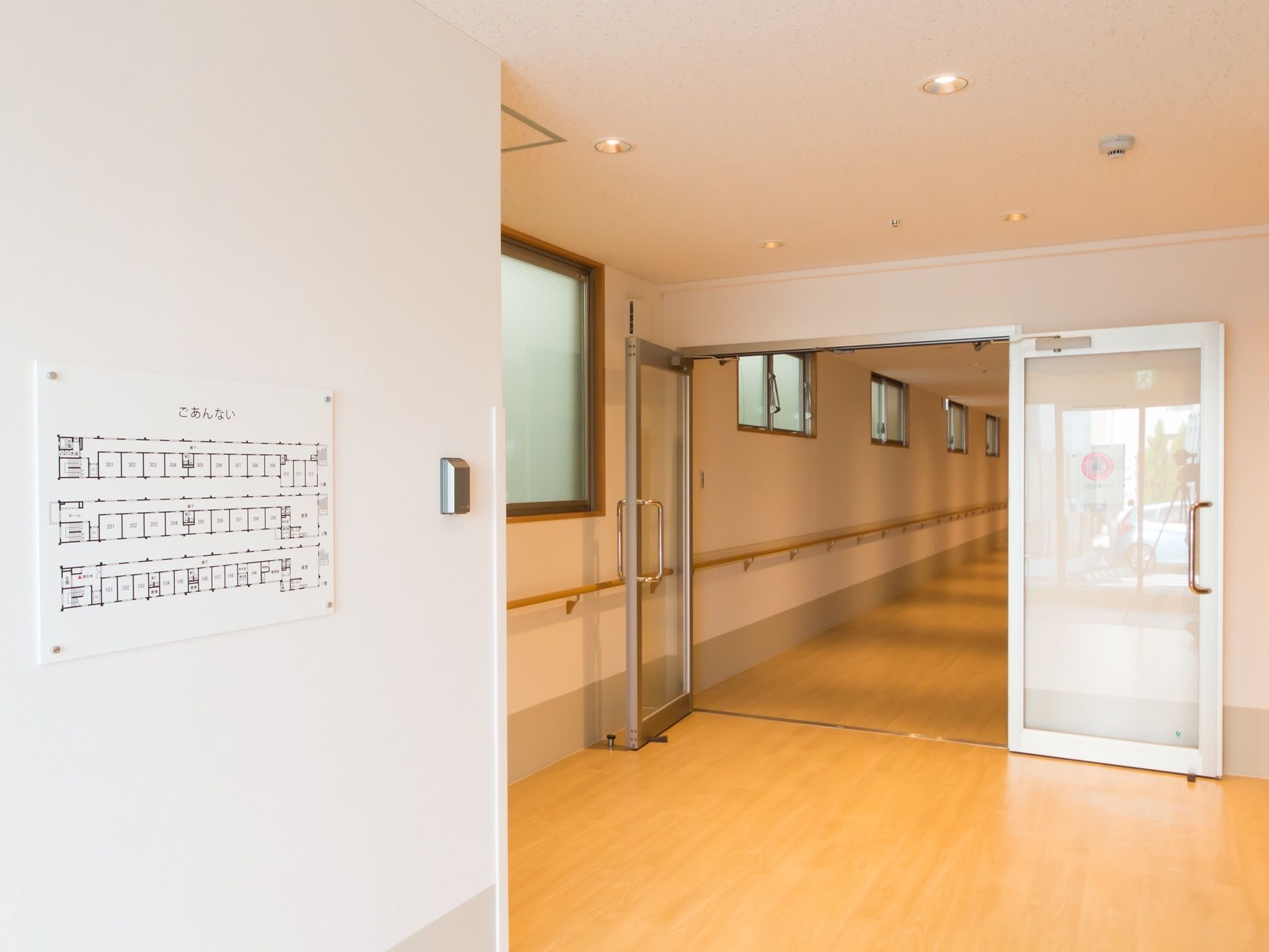 木目調の床の廊下である。奥に進む手前に、両開きのガラス張りの扉がある。壁には、敷地内の案内図を貼っている。