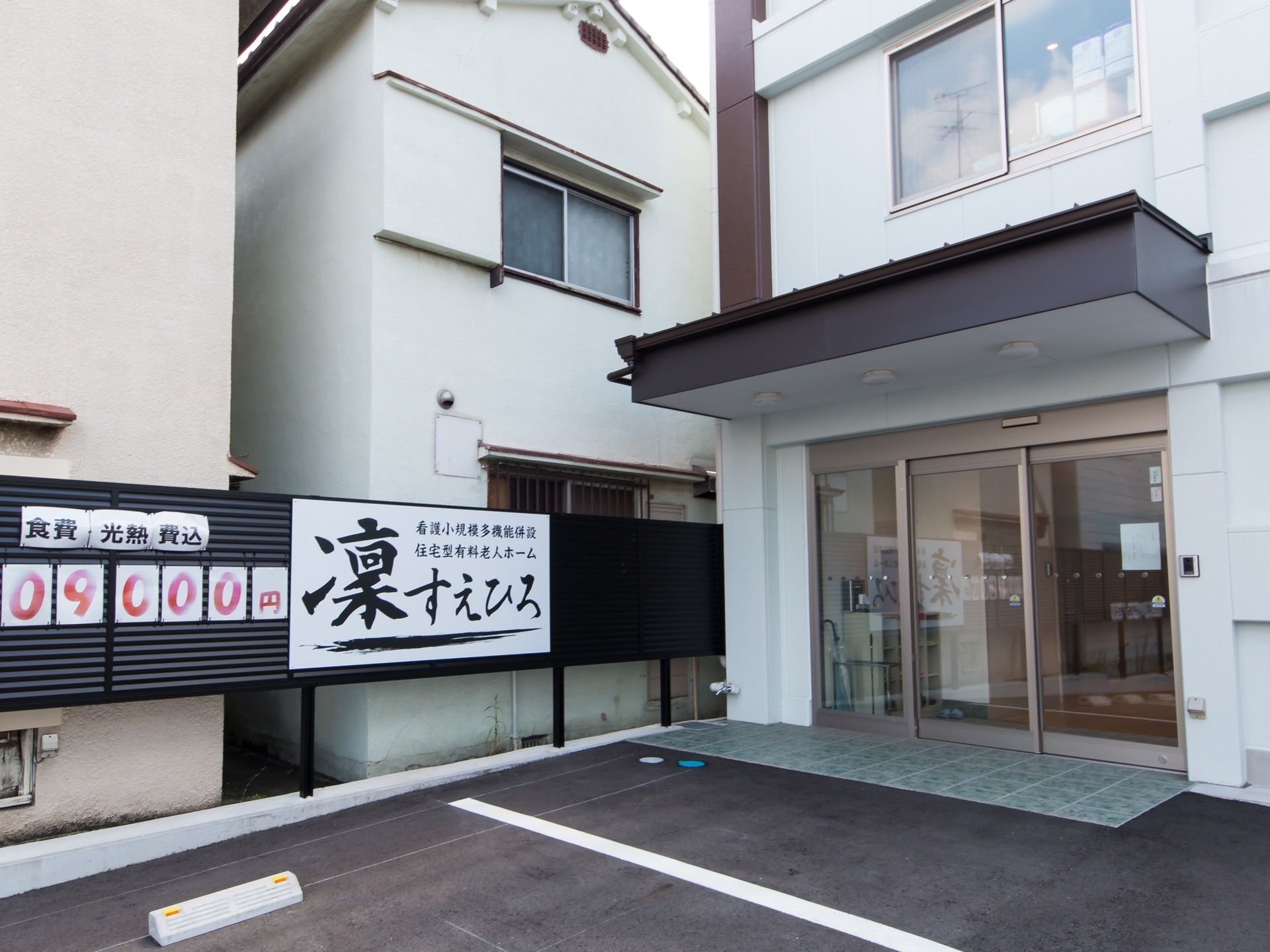 ガラス張りの自動ドアの玄関である。横側に柵を設置し、大きな施設の看板、隣に料金を掲げている。駐車スペースを設けている。