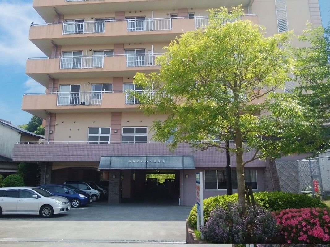 ブラウン系の建物の玄関には事業所が書かれた屋根が付いていて、駐車場には車が止まっている。