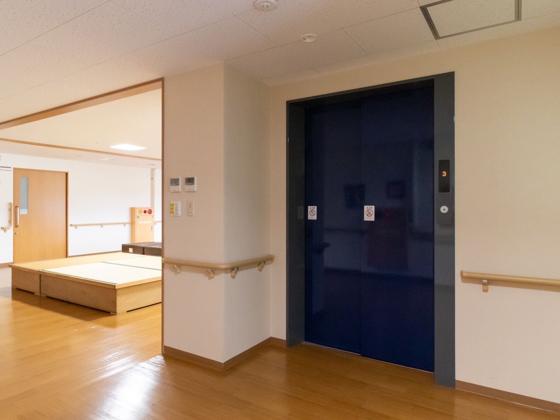 紺色の扉のエレベーターが１基設置されている。扉には正面の様子が写っている。壁には手すりが取り付けられている。