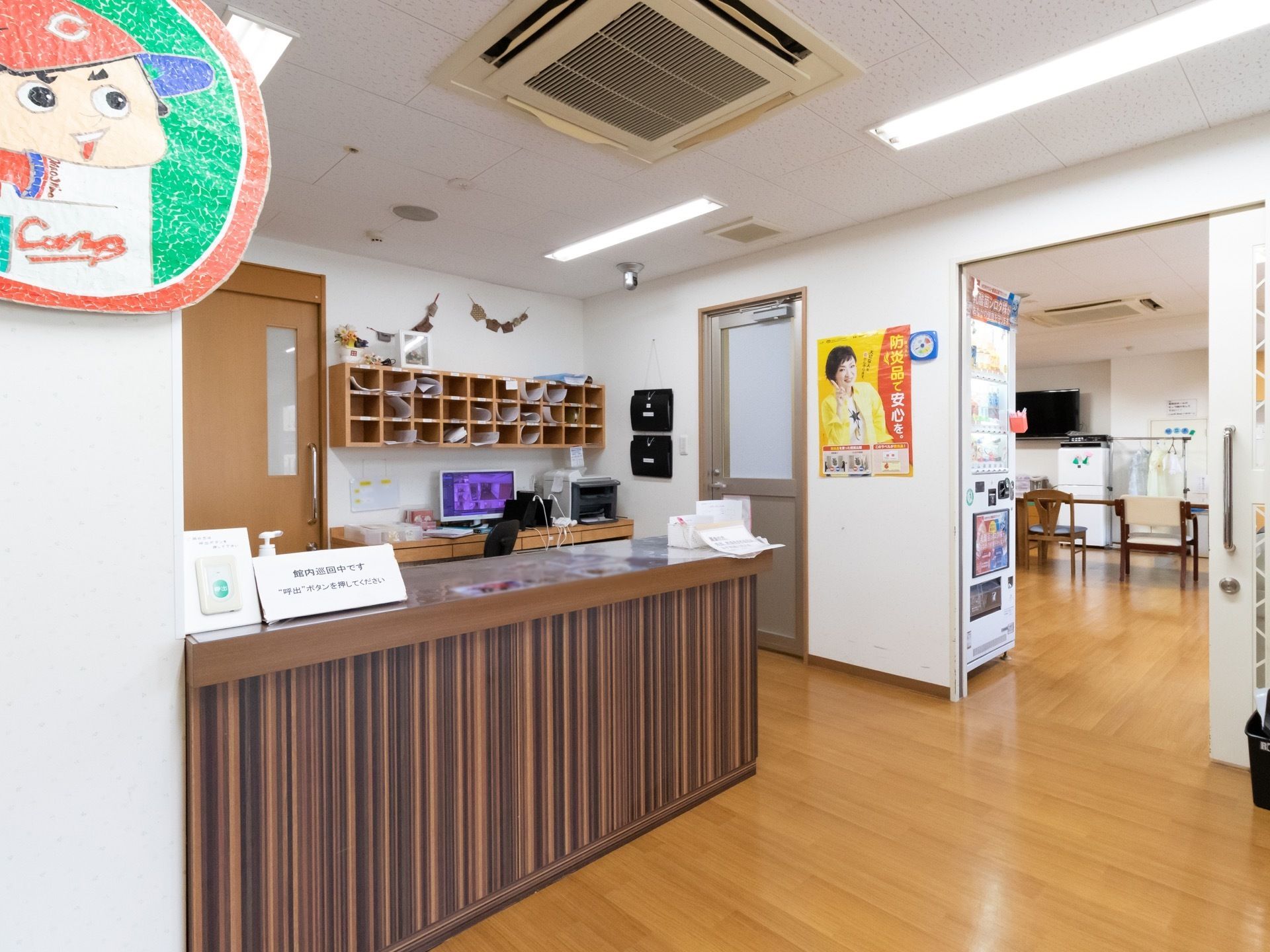 焦げ茶色のカウンターがあり、消毒液と呼び出しボタンが置かれている。横の壁には野球チームのマスコットが飾られている。