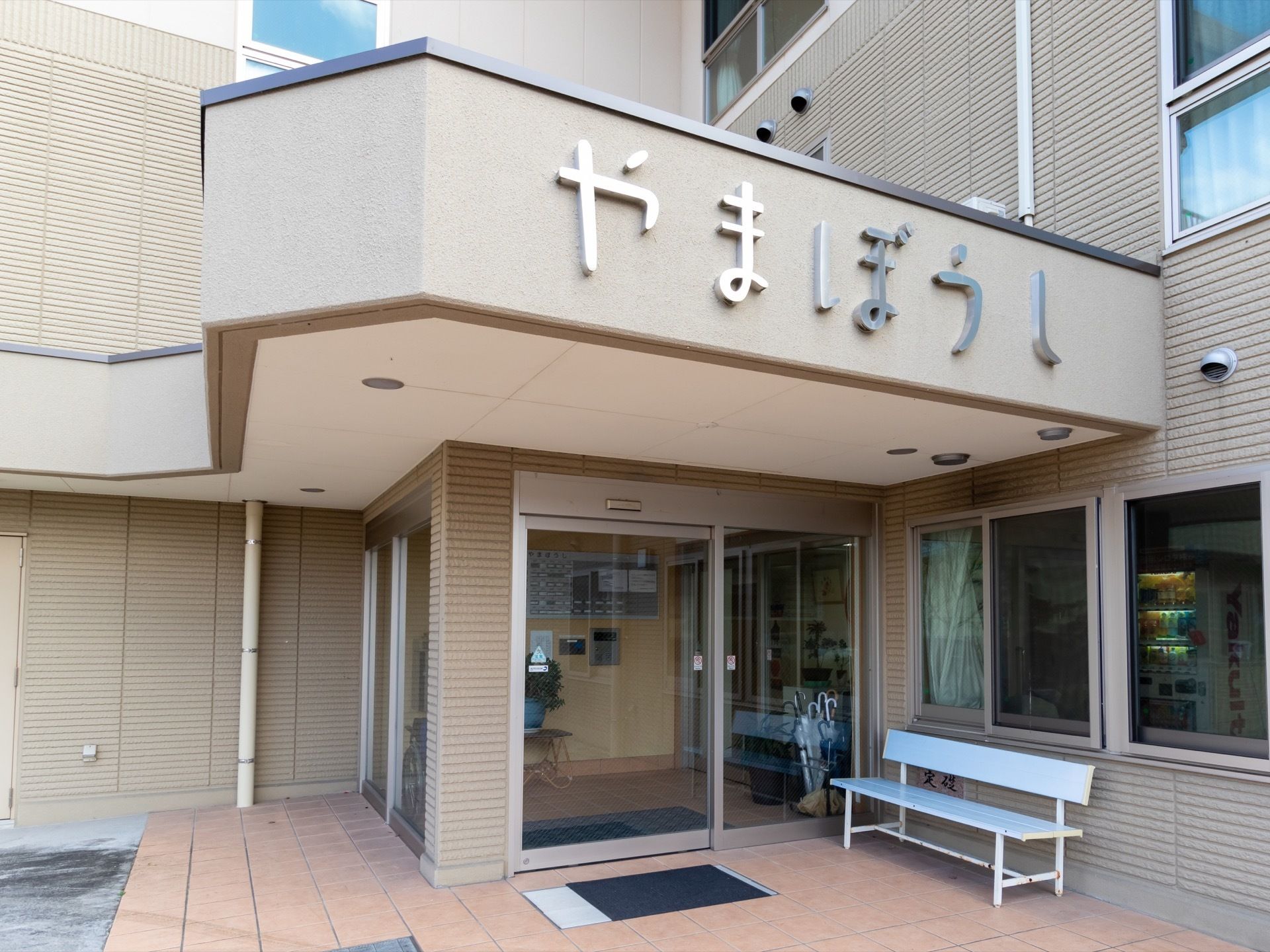 施設玄関の屋根部分の壁に、施設名が取り付けられている。ドアの前には水色のベンチが置かれているほか、マットが敷いてある。