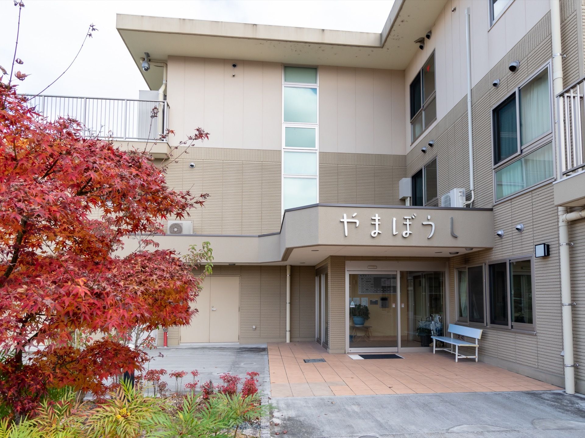 ３階建ての建物の正面から見た様子である。玄関の上には、施設名が大きく書かれており、ドアの前にベンチが置いてある。