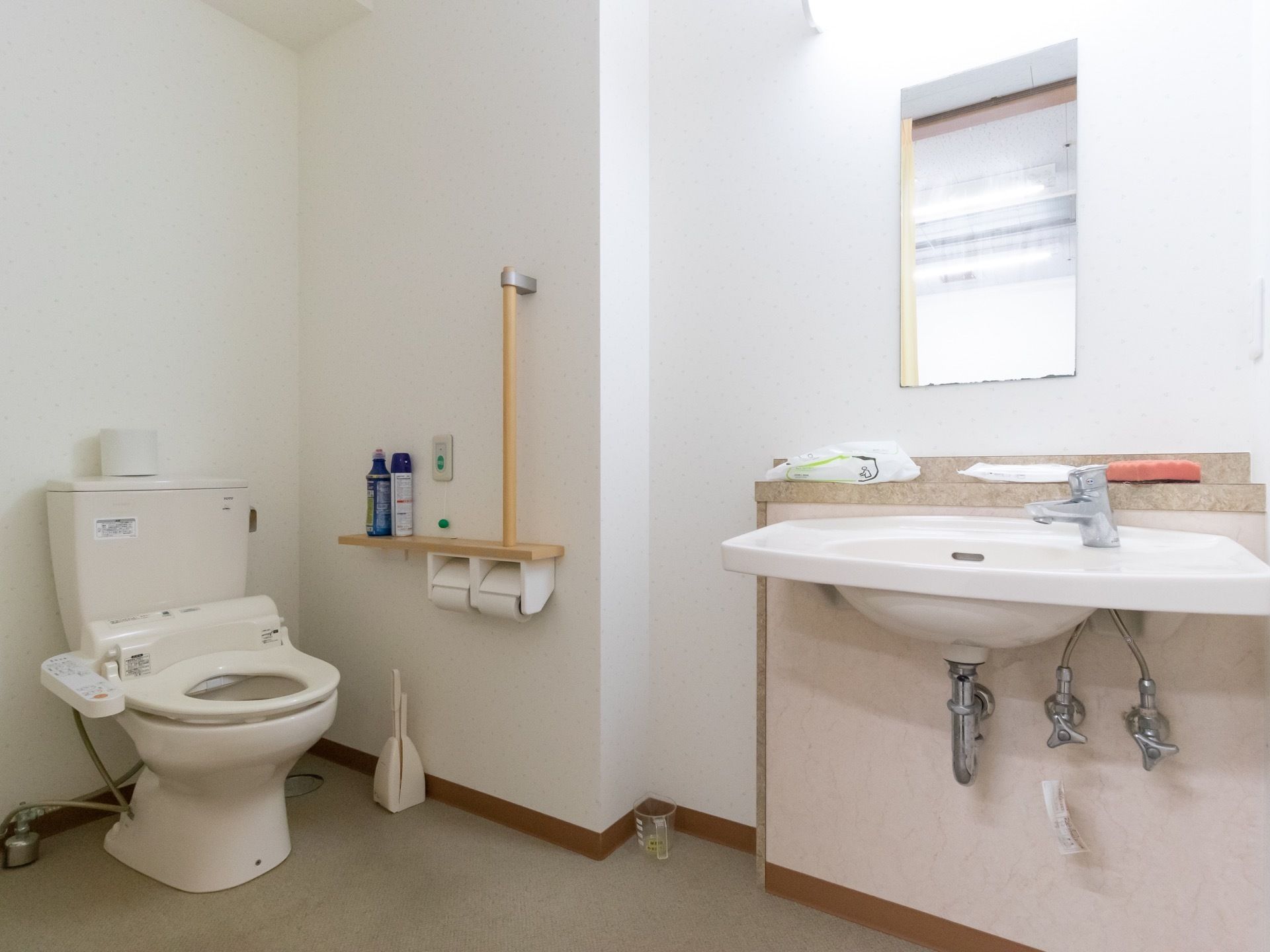 鏡付きの手洗い場があるトイレである。床には掃除道具が置かれており、トイレ後ろのタンクの上にはトイレットペーパーが１個ある。