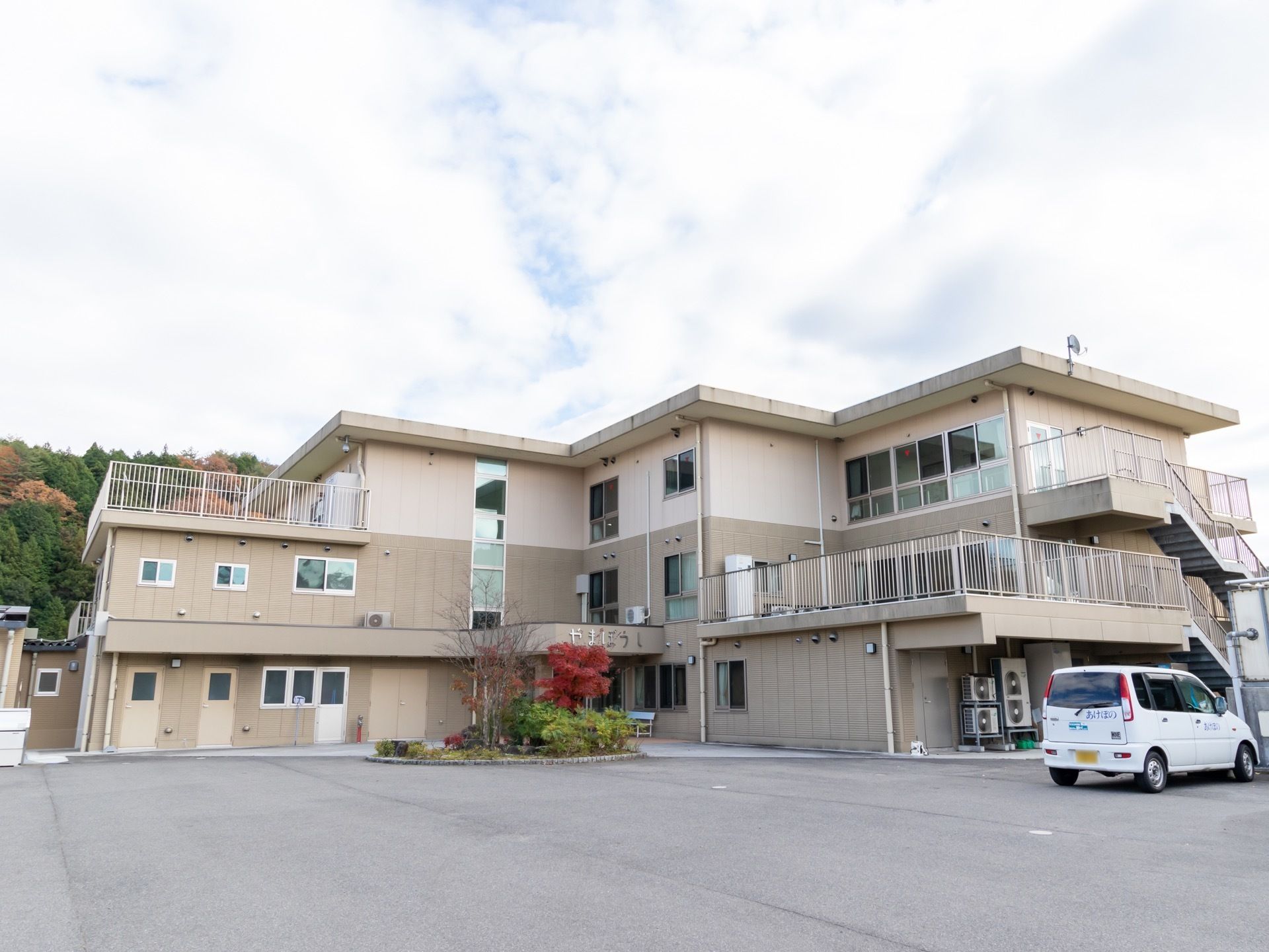 奥に緑の森が見える場所にある、３階建ての建物である。2階と３階の一部分には、ベランダが設置されており、外階段もある。