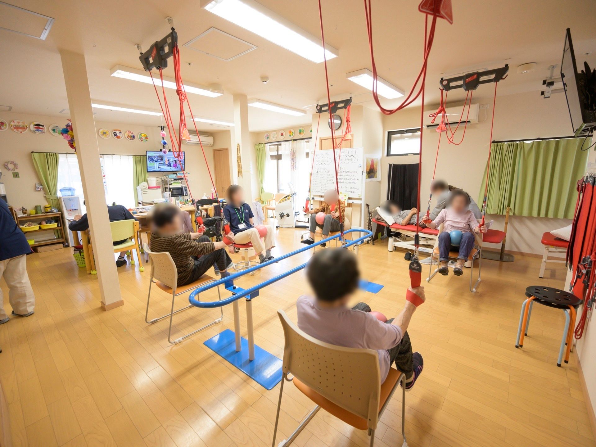 フローリングのホールに、スリングロープや歩行訓練のバーが設置されている。入居者様が数名、スタッフと共に身体を動かしている。