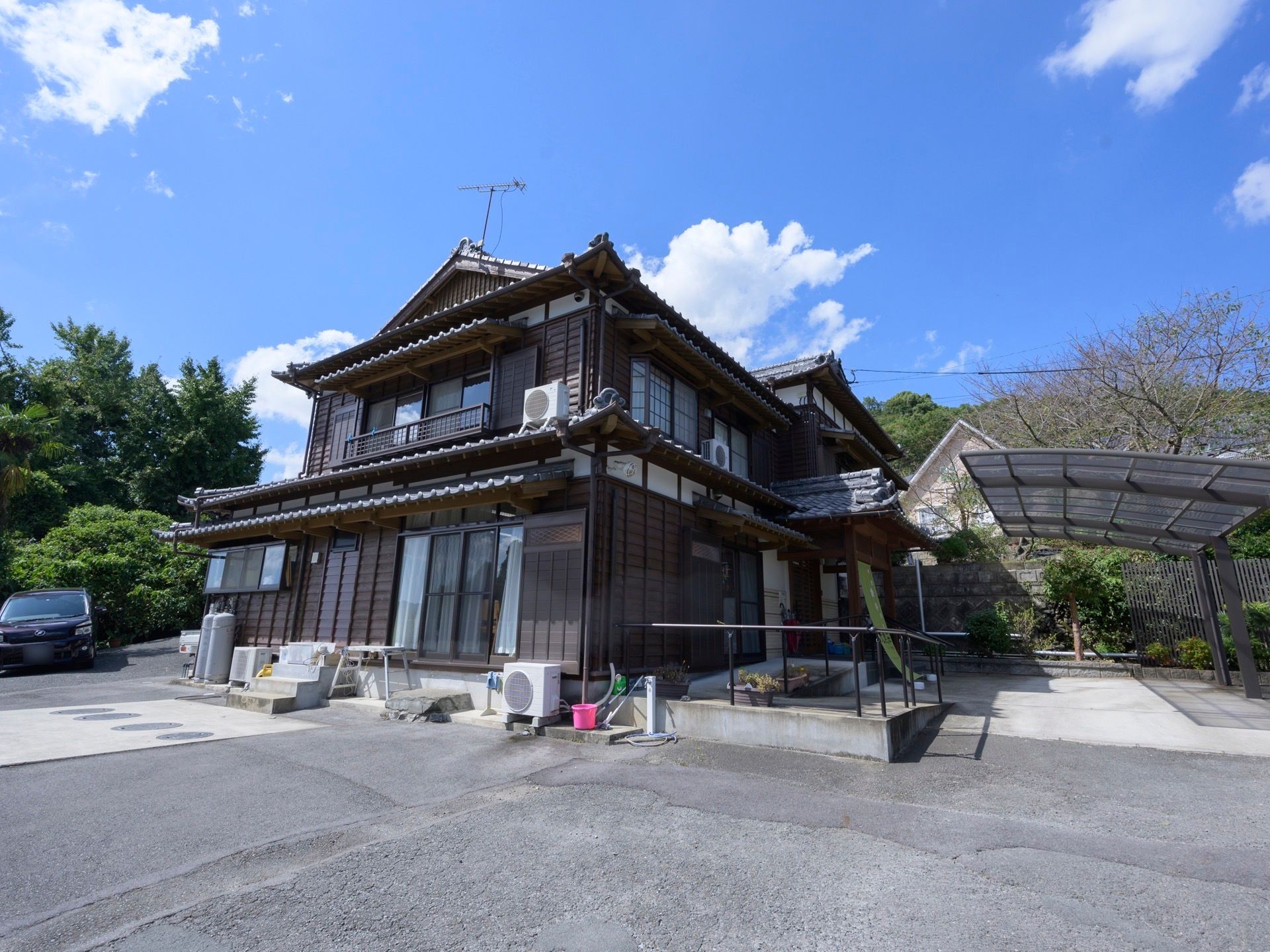 黒い瓦屋根の日本家屋が建っている。玄関前は手すりの付いたスロープがあり、舗装された駐車スペースが用意されている。
