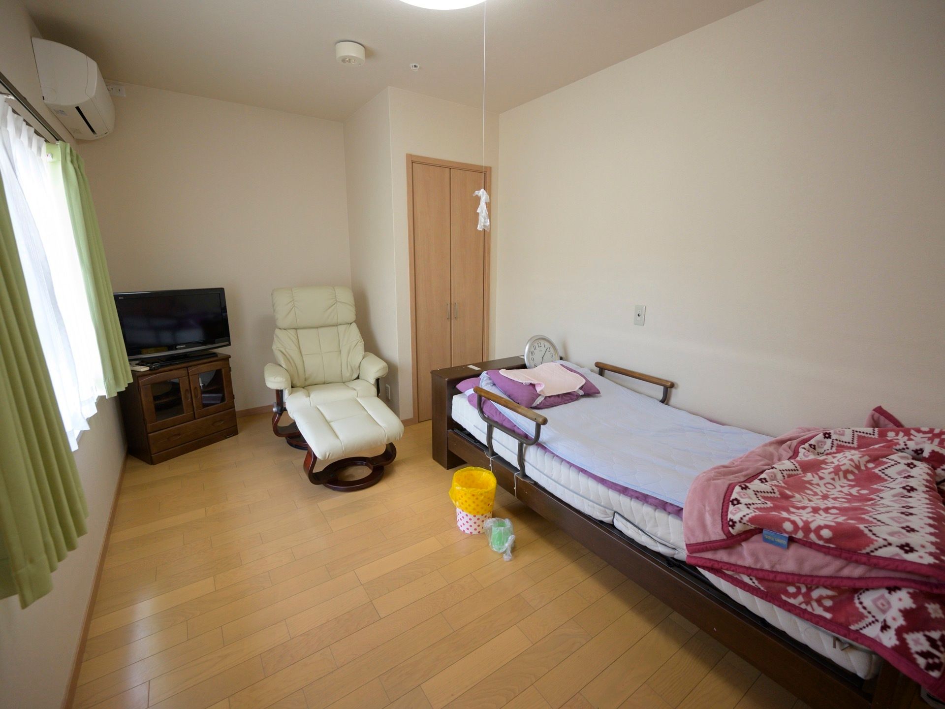 エアコンが付いたフローリングの居室があり、窓の近くにテレビと椅子、反対の壁際には電動ベッドが１台置かれている。