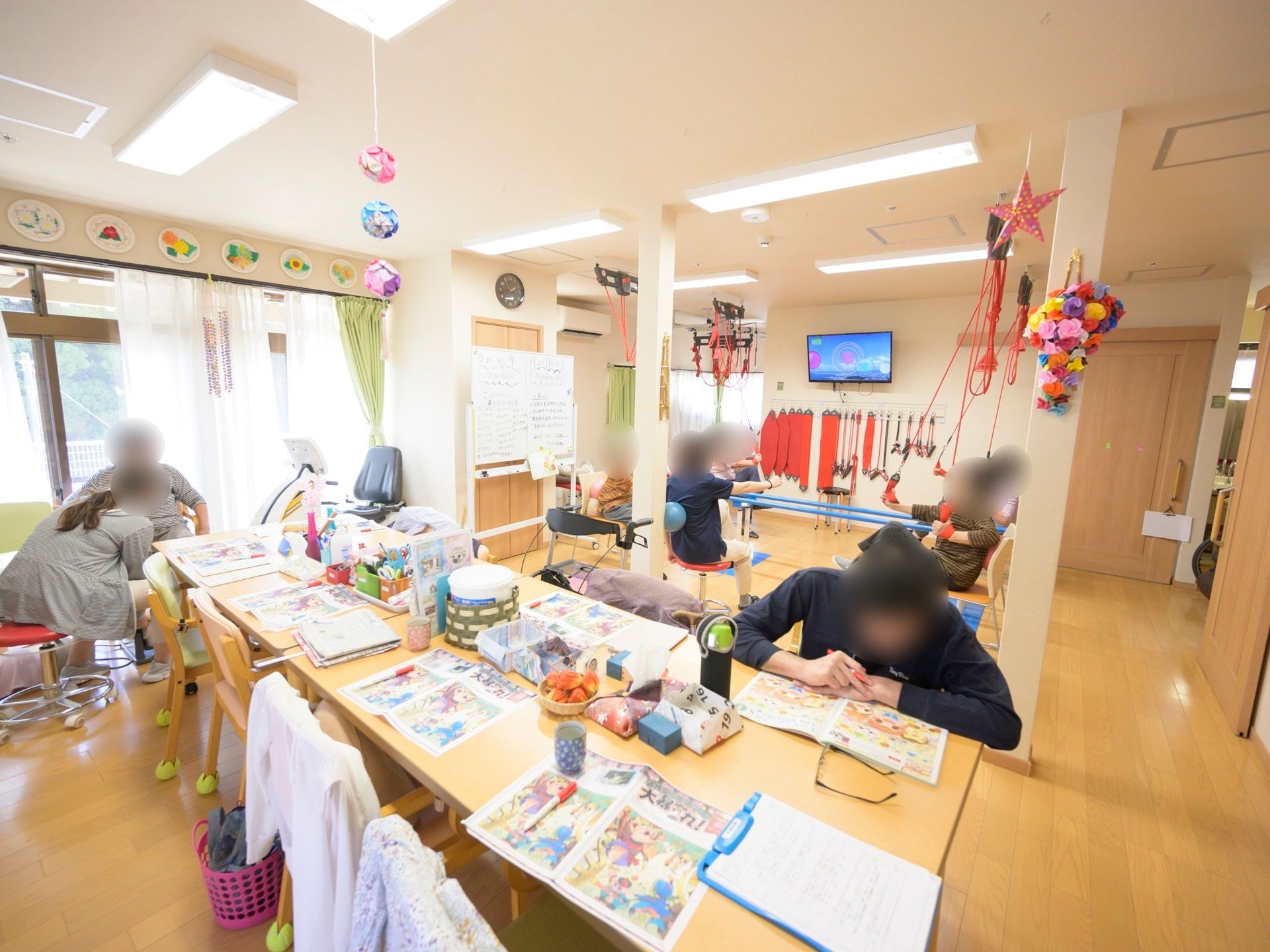 ダイニングテーブルが一列に並べられたフロアがあり、奥にはトレーニング機器が置かれている。入居者様がスタッフと共に身体を動かしている。