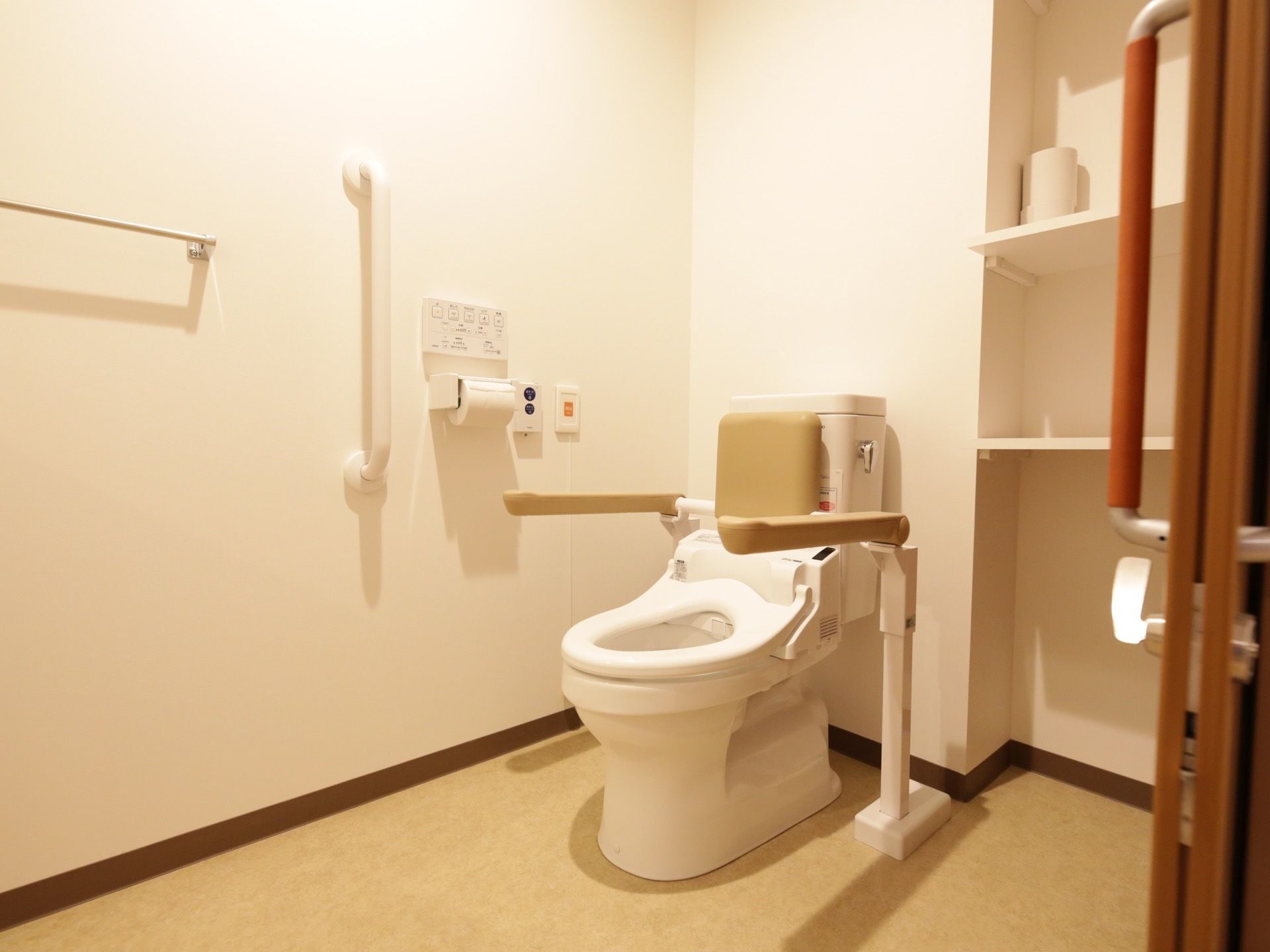 白い壁の広いトイレがある。便座にはひじ掛け、壁には手すりがある。側には備え付けの大きな棚がある。