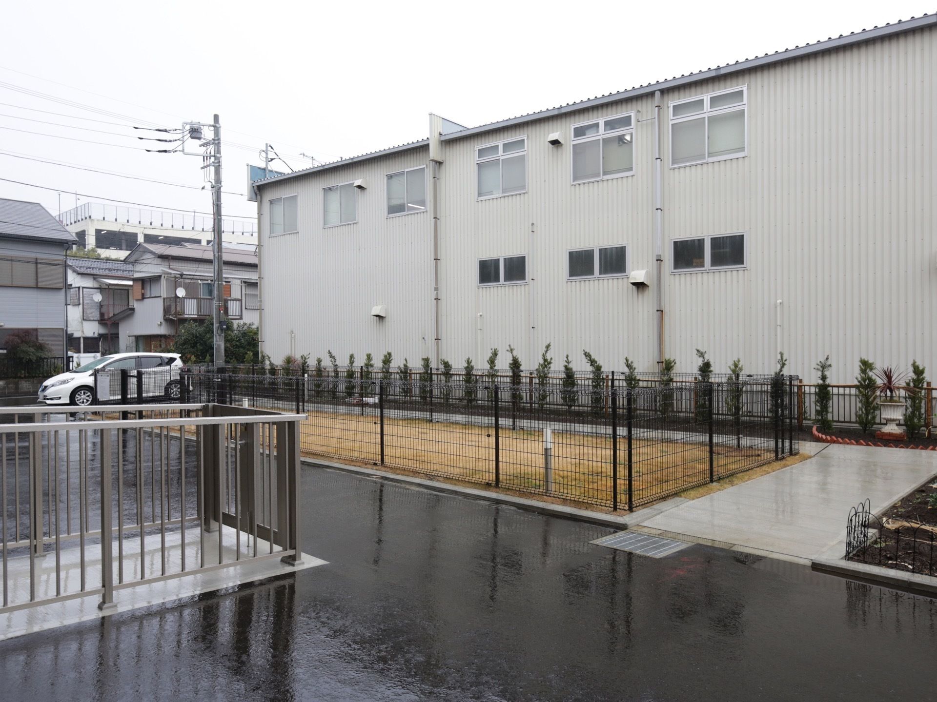 建物の裏側に通路がある。地面は舗装され、フェンスで仕切られた芝生の繁ったスペースがある。敷地の周りには柵があり、樹木が植えられている。